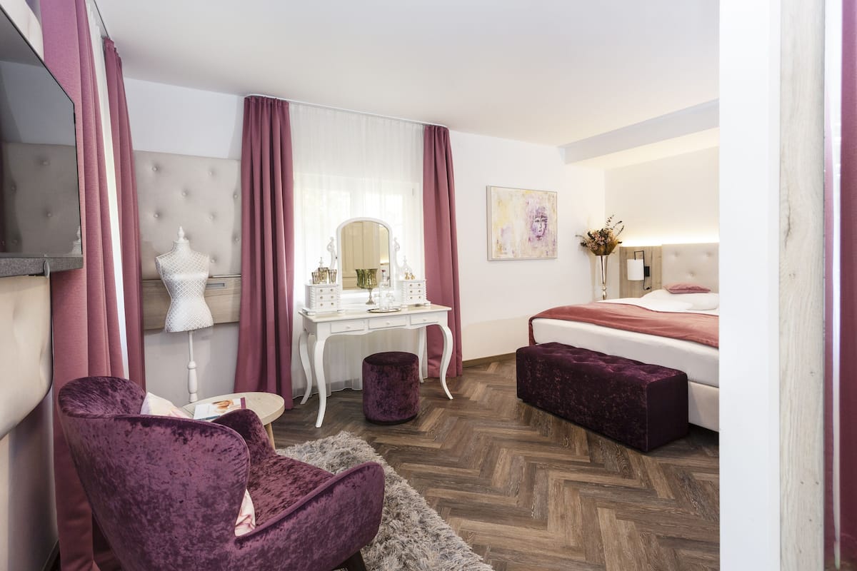 A hotel room with a bed, a vanity with a mirror, a chair, a small table, and a decorative plant.
