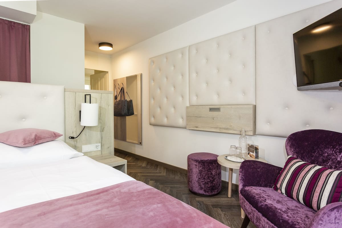 A modern hotel room with a large bed, a tufted headboard, a flat-screen TV mounted on the wall, a purple armchair, and a small round table with a glass.