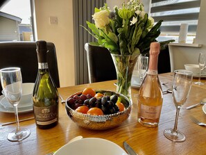A dining table with a bottle of Prosecco, a bowl of fruit, and a vase of flowers. - Four Corners Holiday Home with Hot Tub & Sauna (Ferndown)