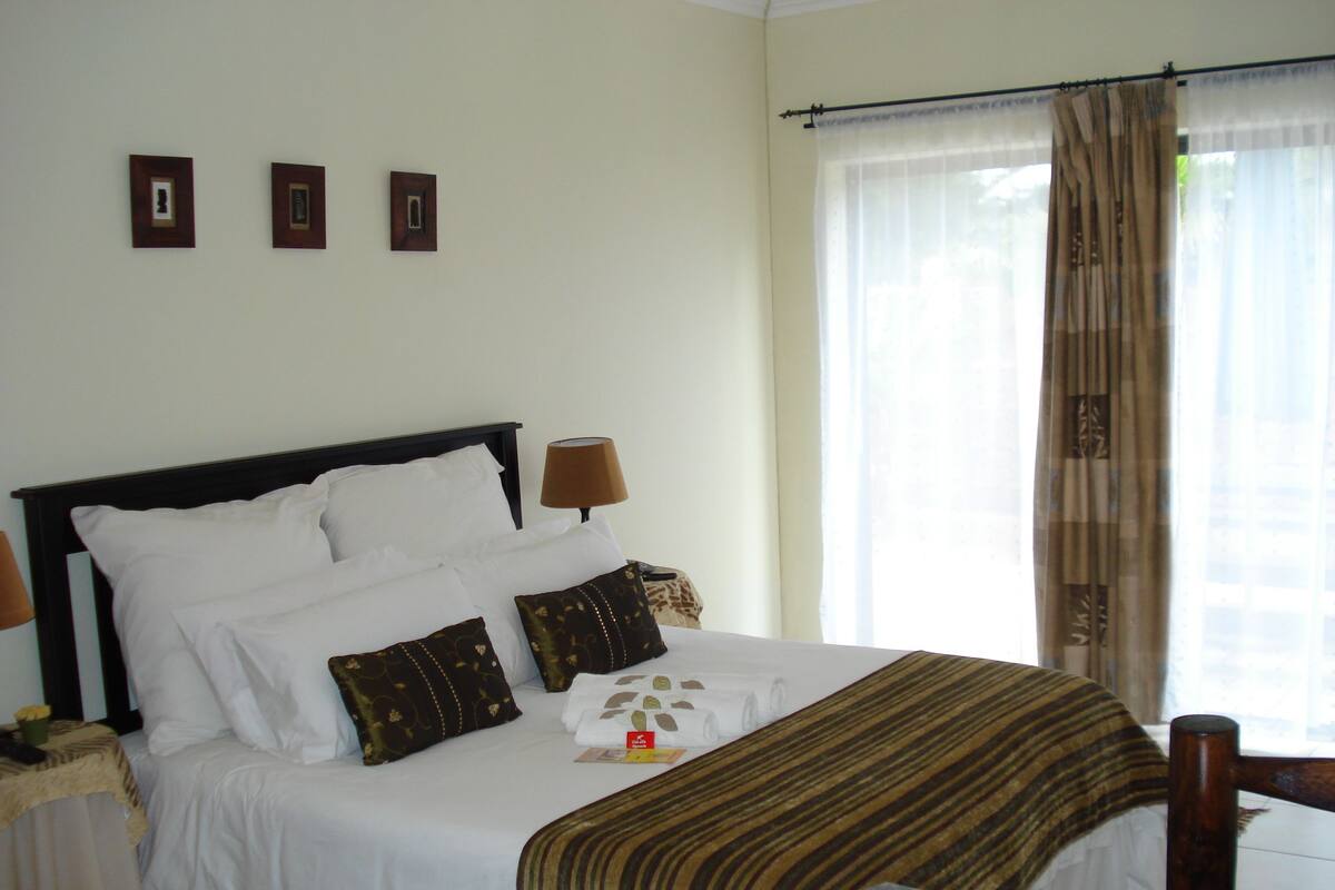A neatly made bed with white linens and multiple pillows, a bedside table with a lamp, and a window with sheer curtains.