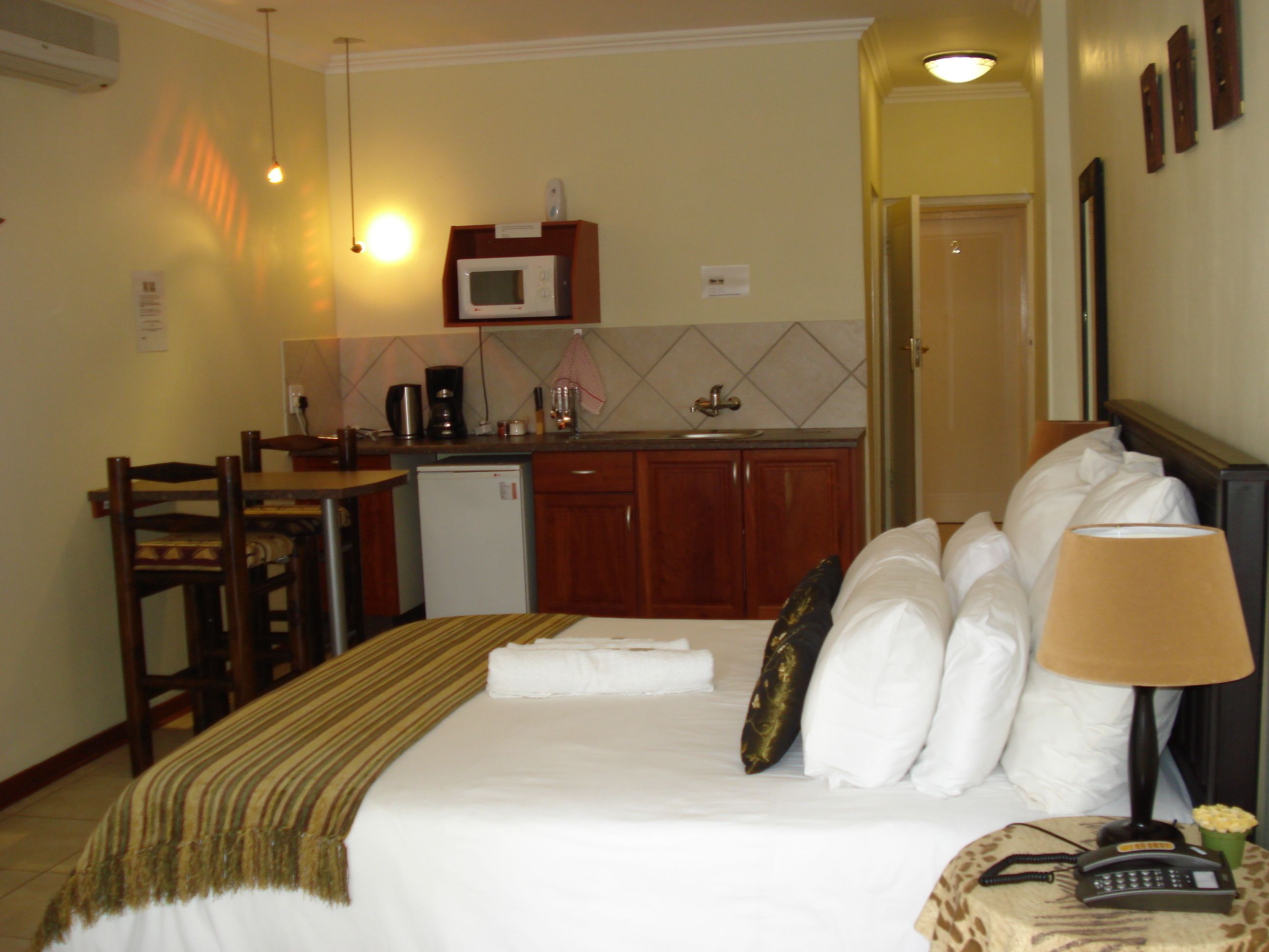 A hotel room with a bed, bedside table, lamp, and a small kitchenette with a microwave, coffee maker, and sink.