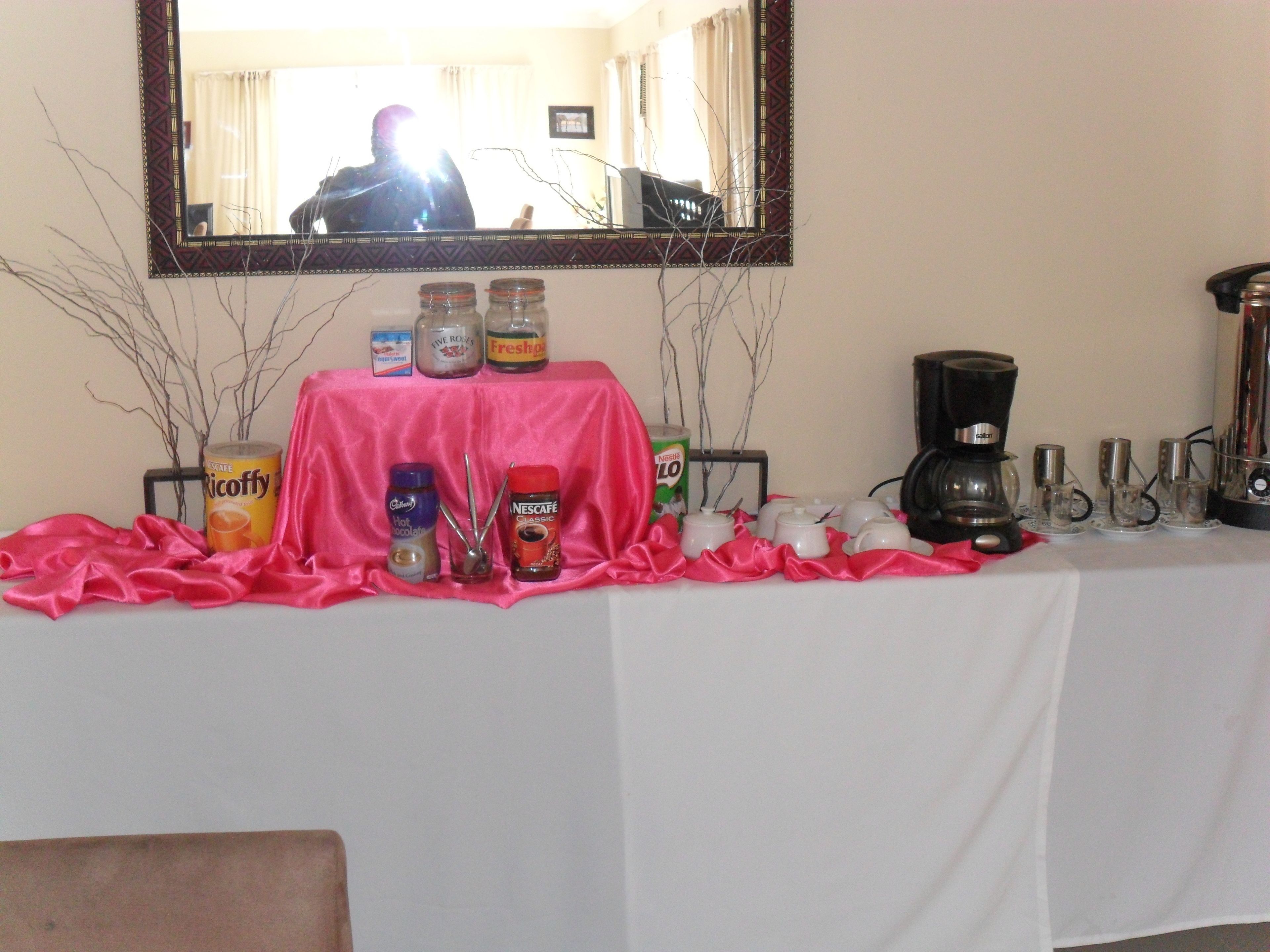 Una mesa con mantel rosa, diversos alimentos y bebidas, una cafetera y un espejo en la pared.