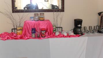 A table with a pink cloth, various food and drink items, a coffee maker, and a mirror on the wall.