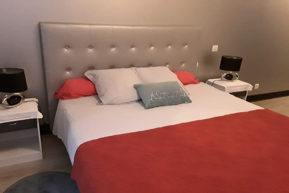 A neatly made bed with a grey headboard, white and red bedding, and a grey patterned pillow. There are two bedside tables with lamps and a small table with a speaker.