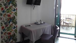 A room with a TV mounted on the wall, a table with a tablecloth, two chairs, and a window with a view of an outdoor area. - Le Madelon (Pays-de-Belves)