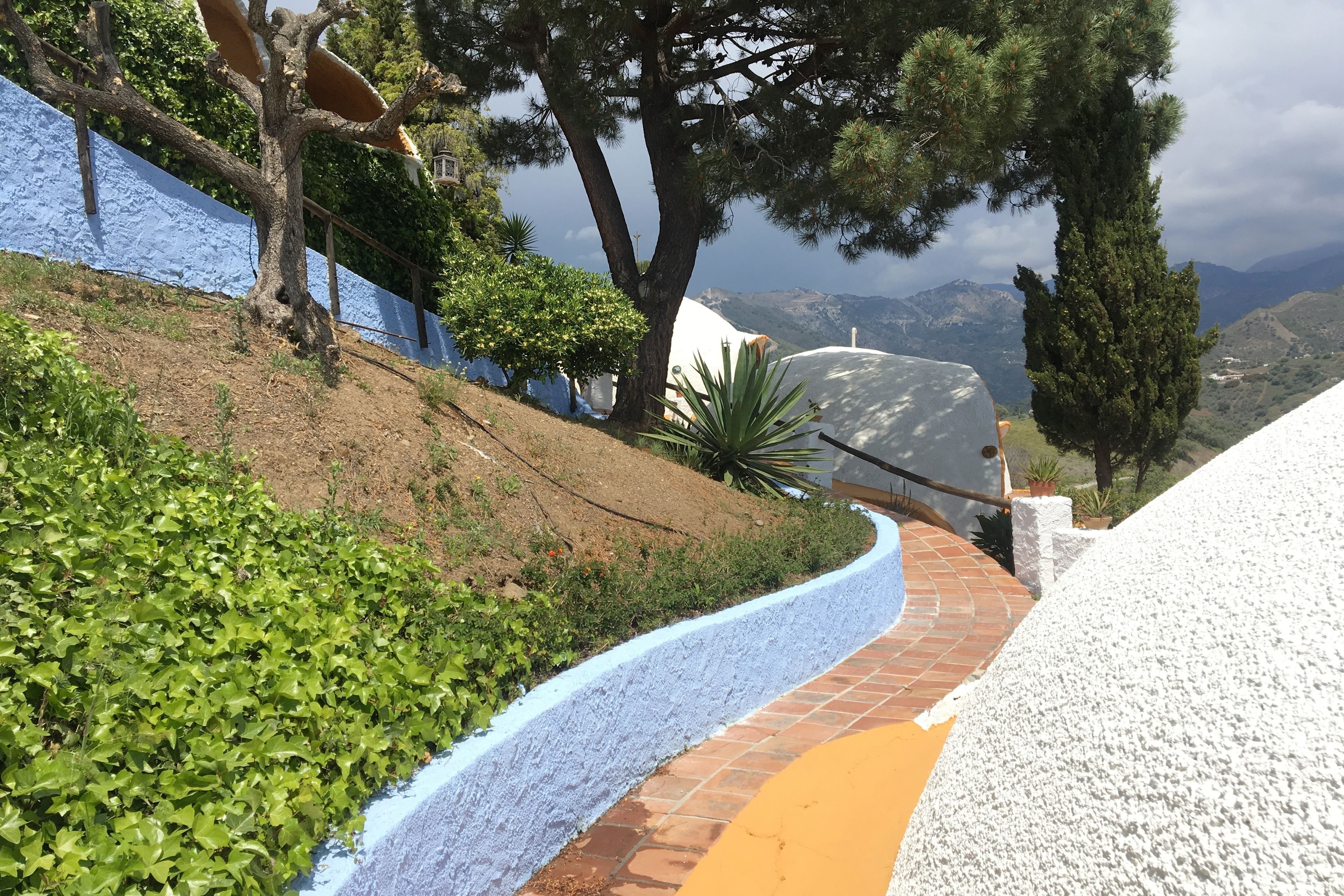 Un sendero serpenteante con un muro azul y blanco a un lado y un camino de ladrillo al otro, rodeado de follaje y árboles.