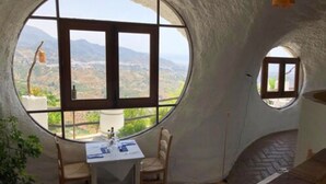 A cozy room with a large circular window offering a scenic view of mountains and greenery. There is a small table with chairs and a lamp. - Hotel Rural Los Caracoles (Torrox)