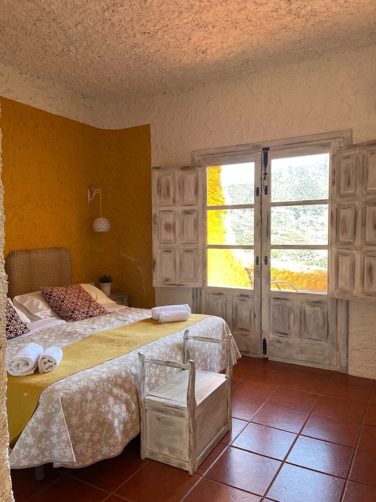 A bedroom with a bed, a bench, a hanging light fixture, and a window with a view of greenery.