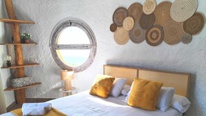 A neatly made bed with white linens, a yellow throw blanket, and rolled towels. There is a round window with a view of the outdoors, a wall-mounted circular woven art piece, and a wooden ladder with shelves holding decorative items. - Hotel Rural Los Caracoles (Torrox)