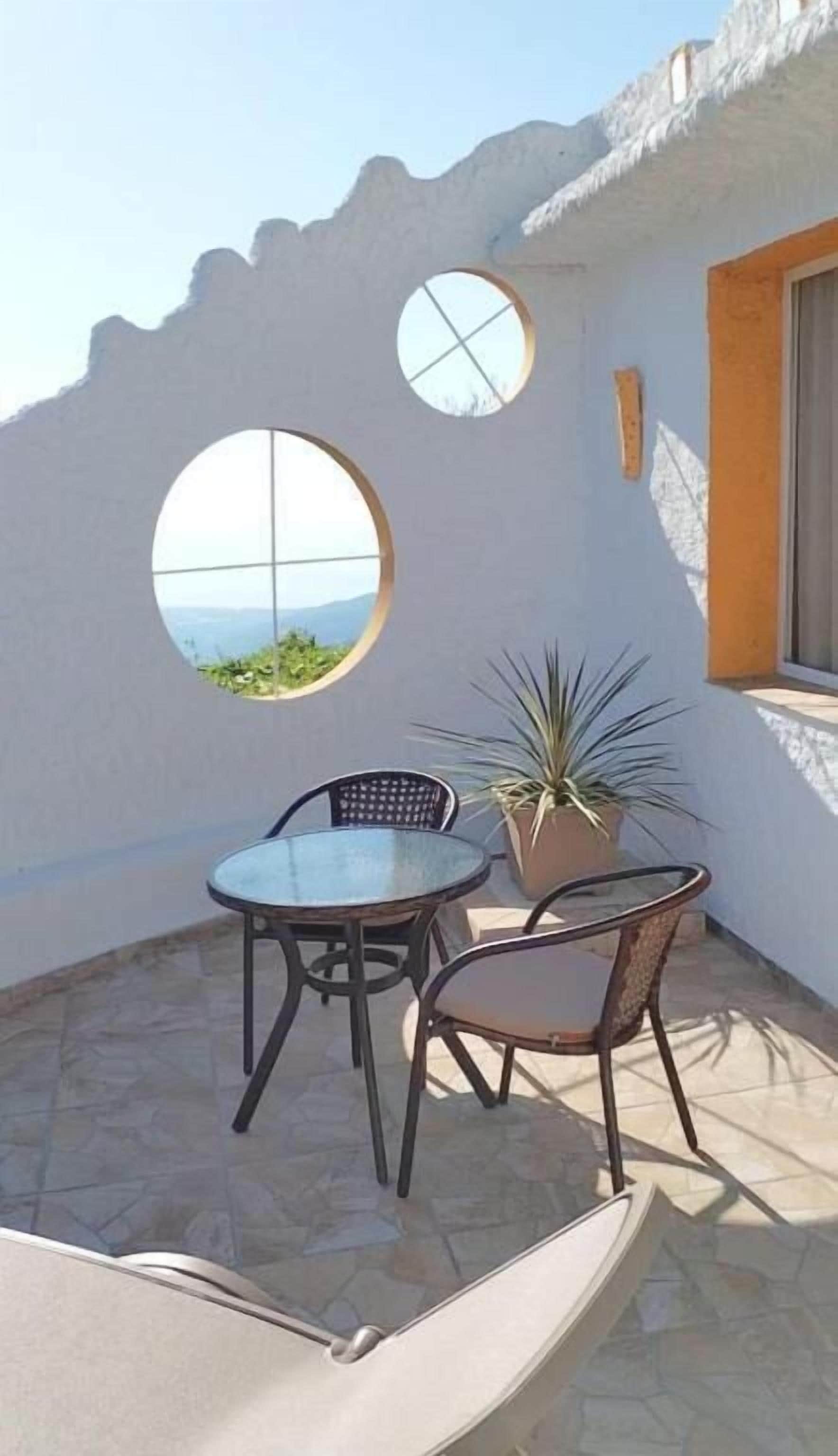 Un pequeño patio con una mesa y dos sillas, una planta en maceta y vista al mar a través de un ventanal circular.