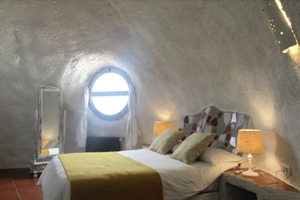 Een slaapkamer met een rond dakvenster, een bed met een gele spreidek, twee bedlampen en een nachtkastje.