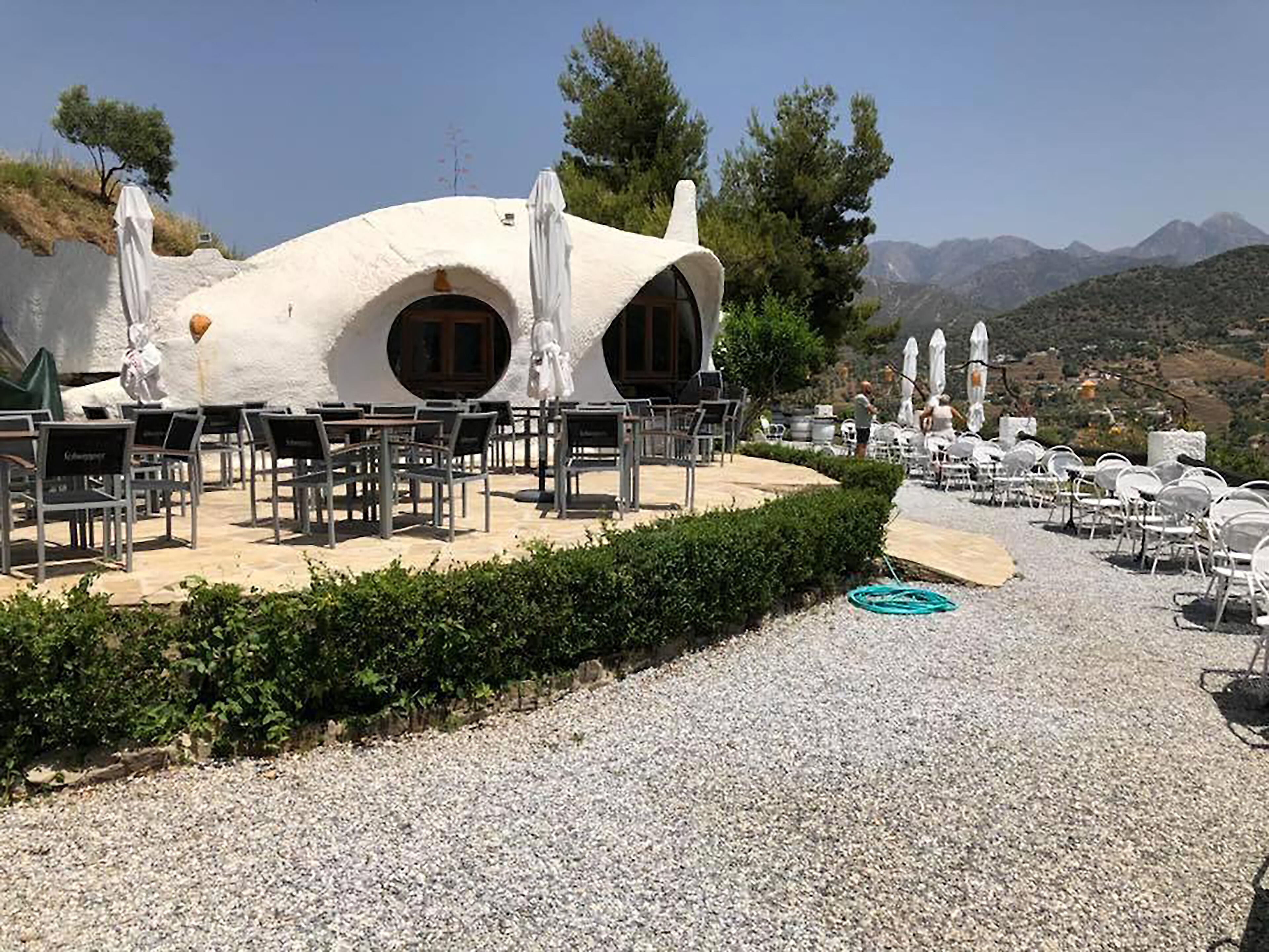 Un edificio blanco de forma abovedada con dos ventanas grandes y un patio de grava con mesas al aire libre.