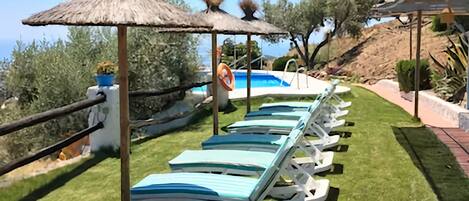 Une rangée de chaises longues, protégées par un parasol en chaume, avec une piscine en arrière-plan.