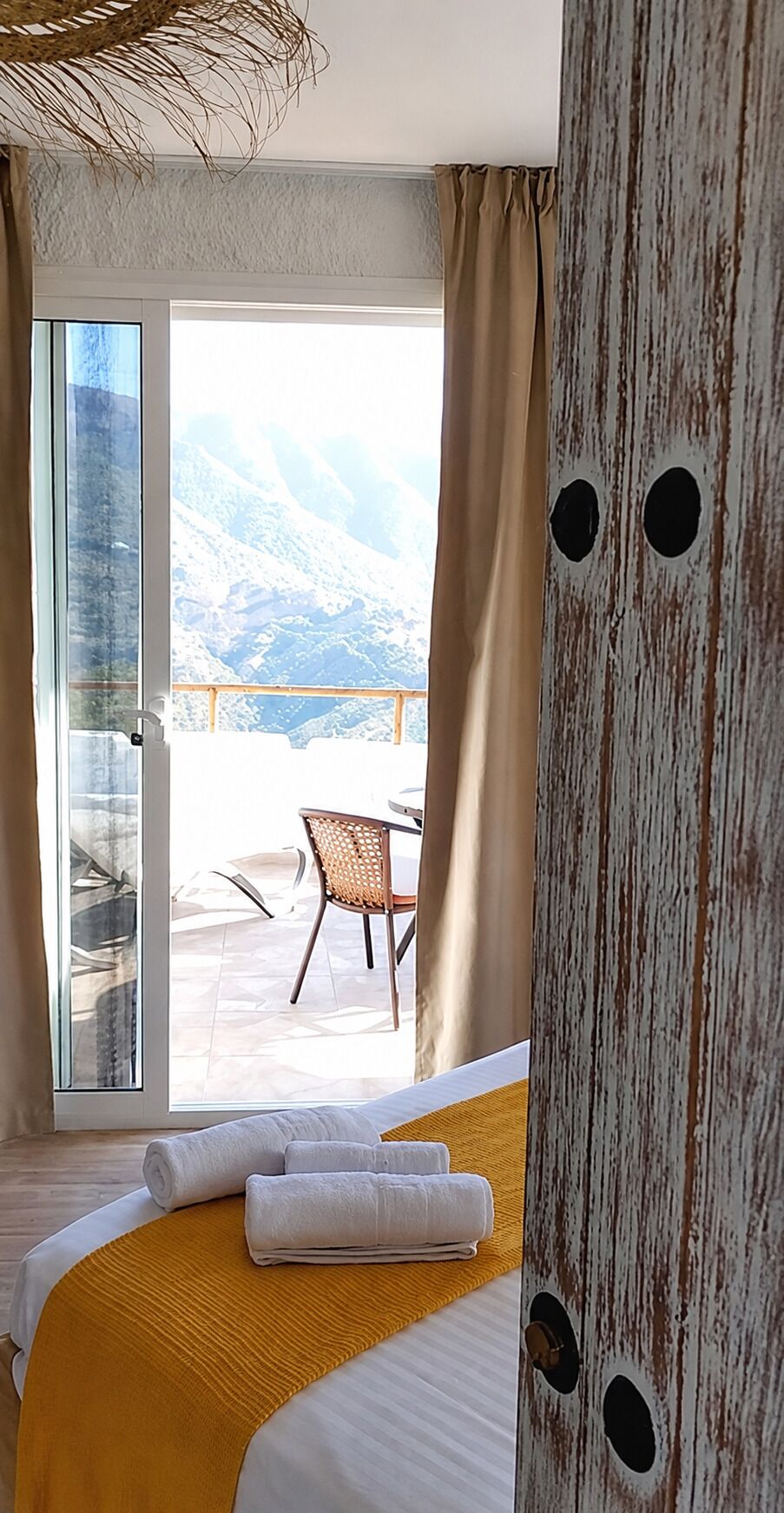 Une chambre d’hôtel avec un lit, une fenêtre donnant sur les montagnes, un balcon avec une chaise et un mur rustique orné d’ouvertures circulaires.