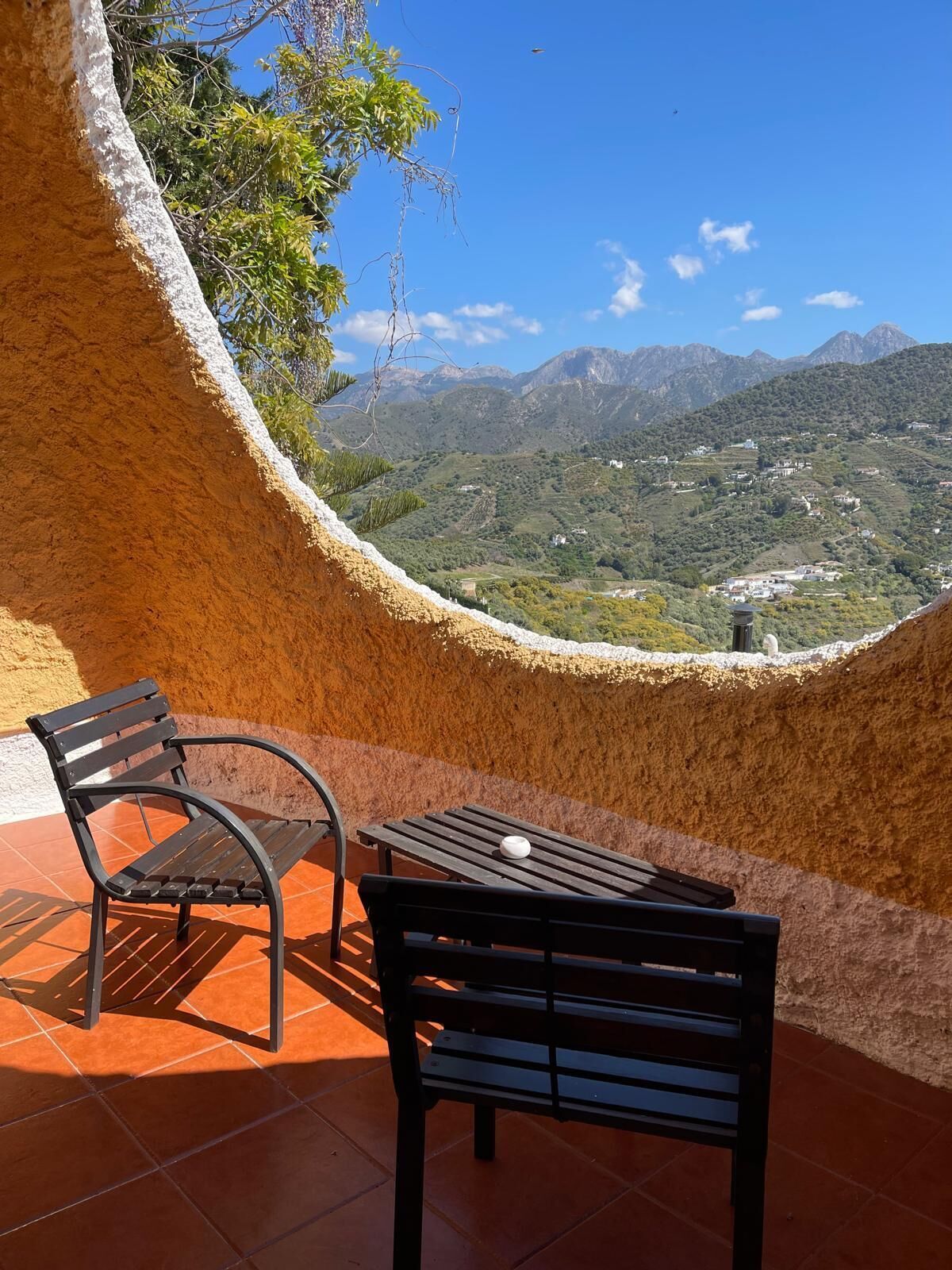 A terrace with two black metal chairs and a table, overlooking a mountainous landscape with buildings.