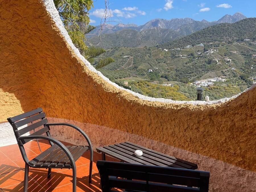 A terrace with two black metal chairs and a table, overlooking a mountainous landscape with buildings.