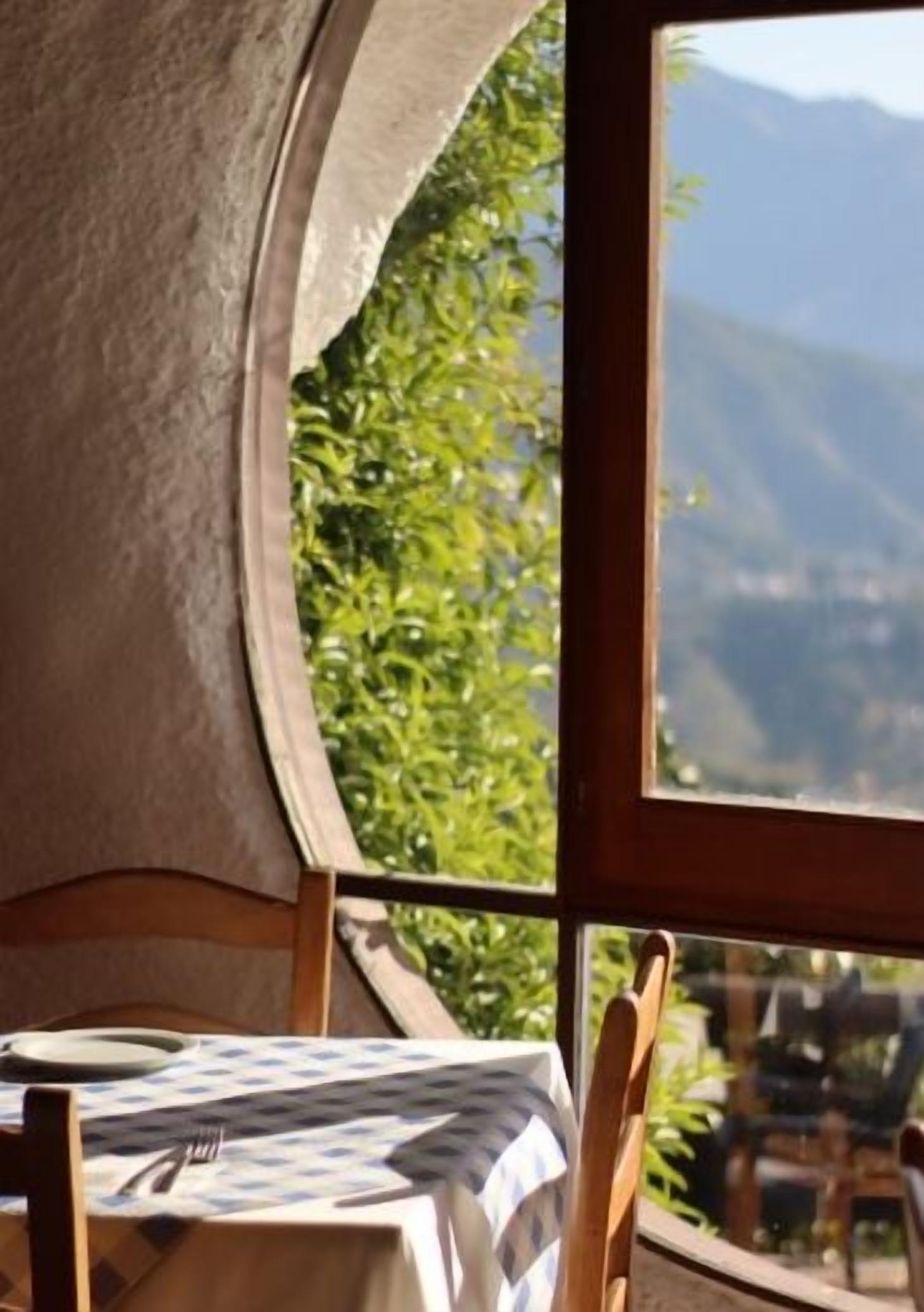 Un coin repas avec une table dressée pour deux personnes, et une vue sur la verdure et les montagnes par la fenêtre.