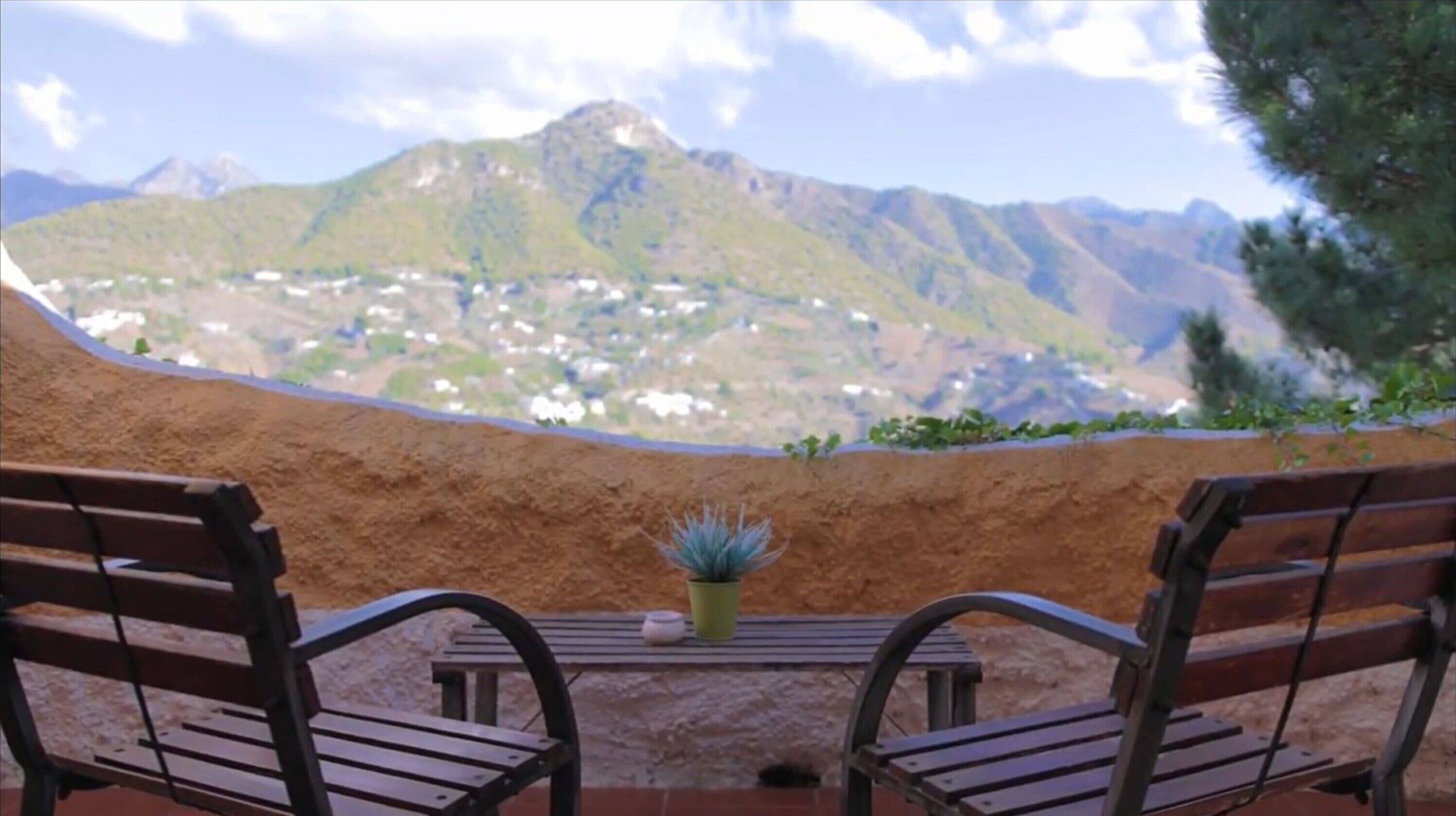 Dos sillas de madera mirando a una mesa con una planta en maceta, con vista a un paisaje montañoso.
