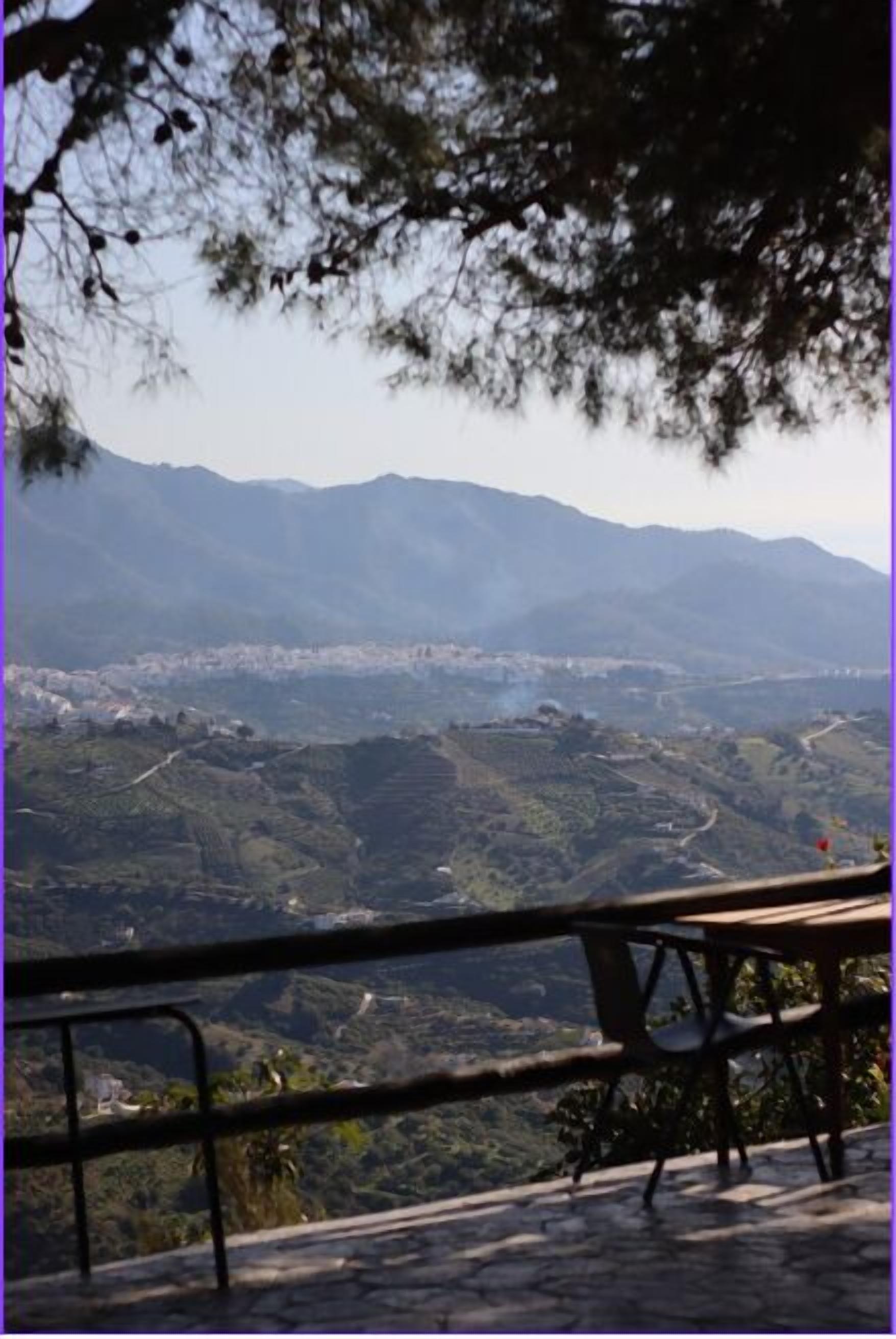 A scenic view from a terrace with a stone floor and metal railing, overlooking a valley with terraced fields and distant mountains.