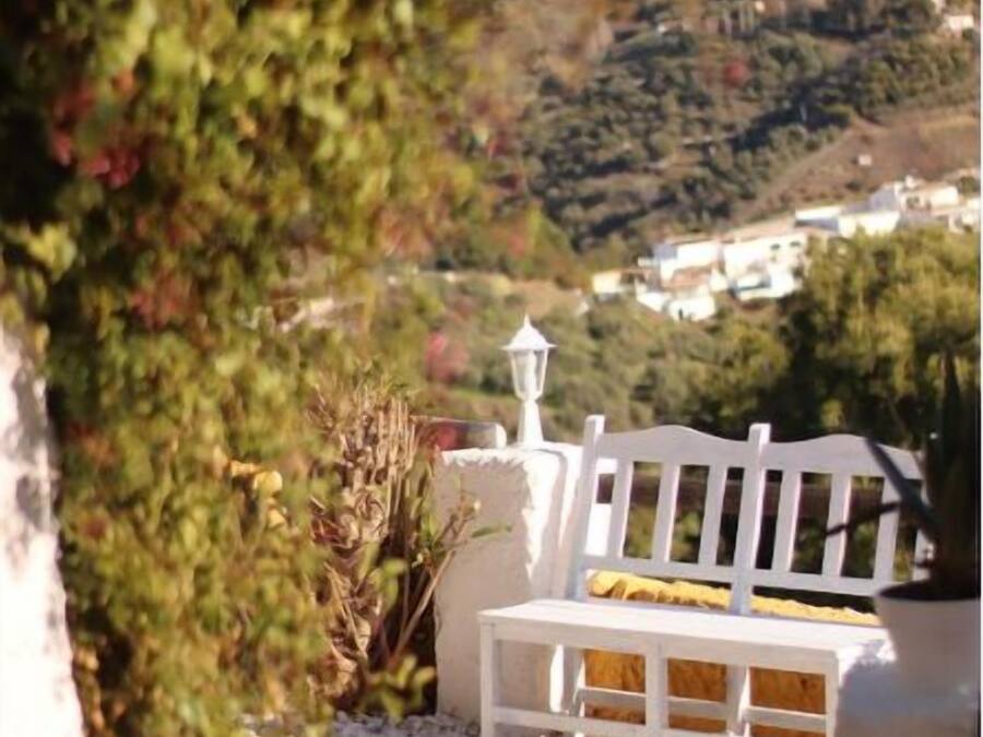 A white bench with a yellow cushion, a white lantern, and a white fence in a garden setting with a view of a hillside.