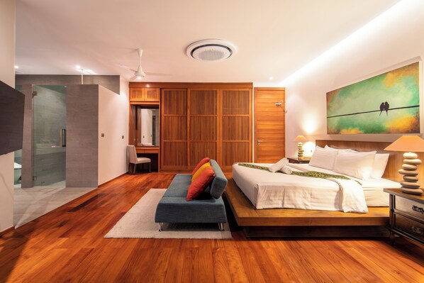 Een moderne slaapkamer met een houten vloer, een bed met witte lakens, een blauwe bank met kleurrijke kussens, een houten kledingkast, en een schilderij van vogels op een draad.