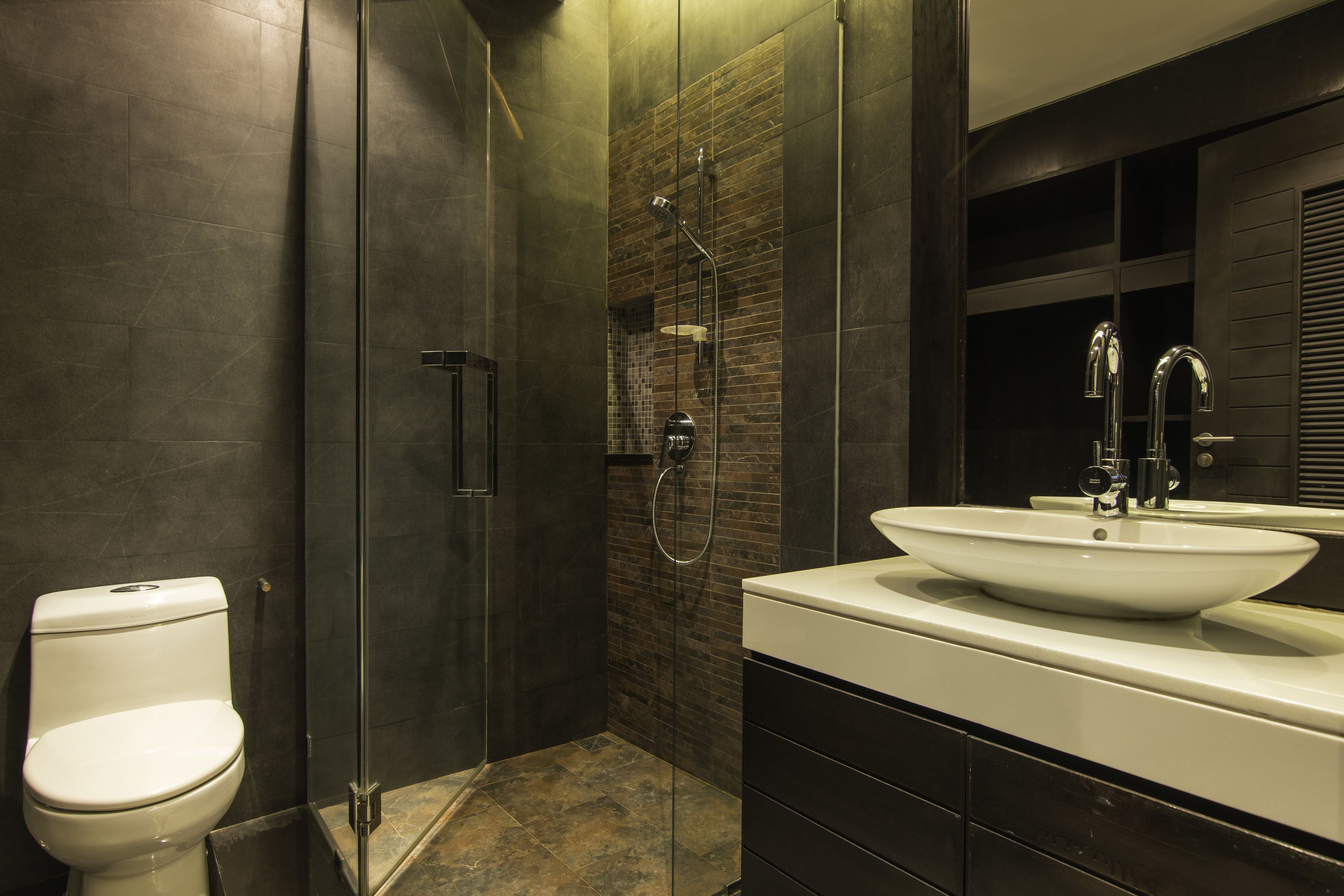 Un baño moderno con ducha de vidrio, lavamanos blanco y azulejos en tonos oscuros.