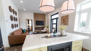A modern kitchen with a central island, a built-in oven, and a microwave. There is a seating area with a sofa and a television in the background.
