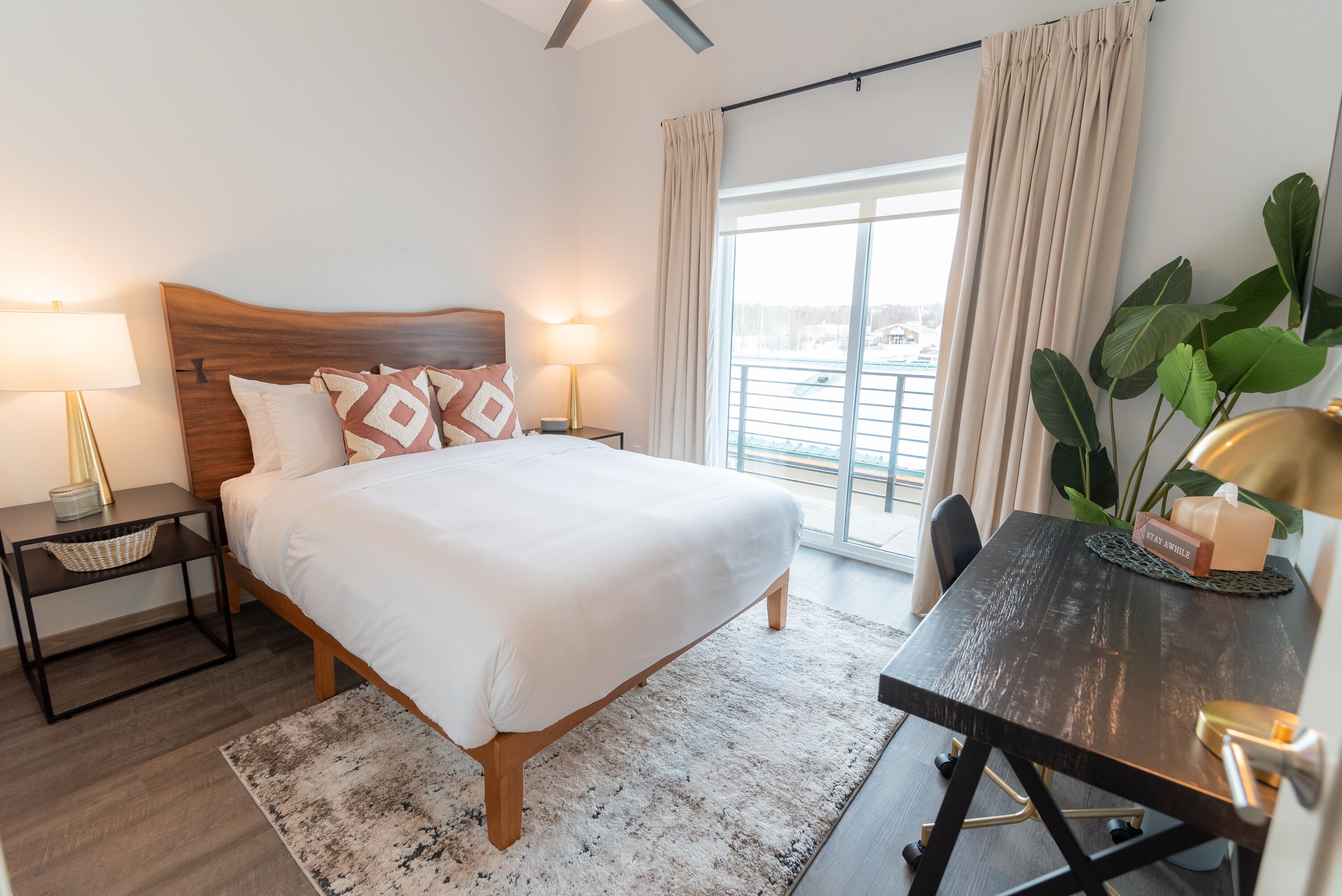 Een hotelkamer met een groot bed, bedlampen, een bureau met stoel, een potplant en uitzicht op het omliggende landschap.
