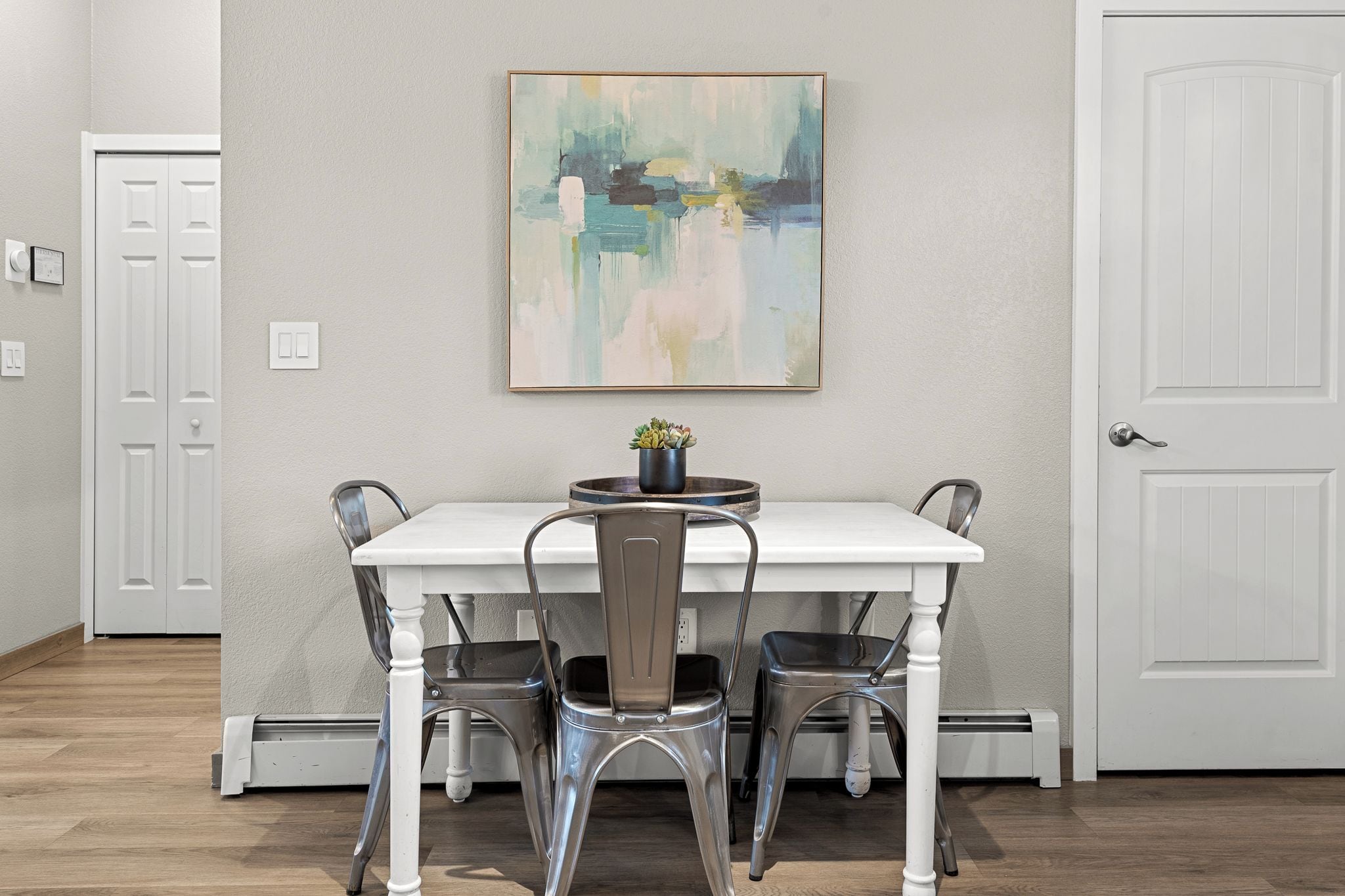 A dining area with a white table and four chairs, a painting on the wall, and a door to the left.