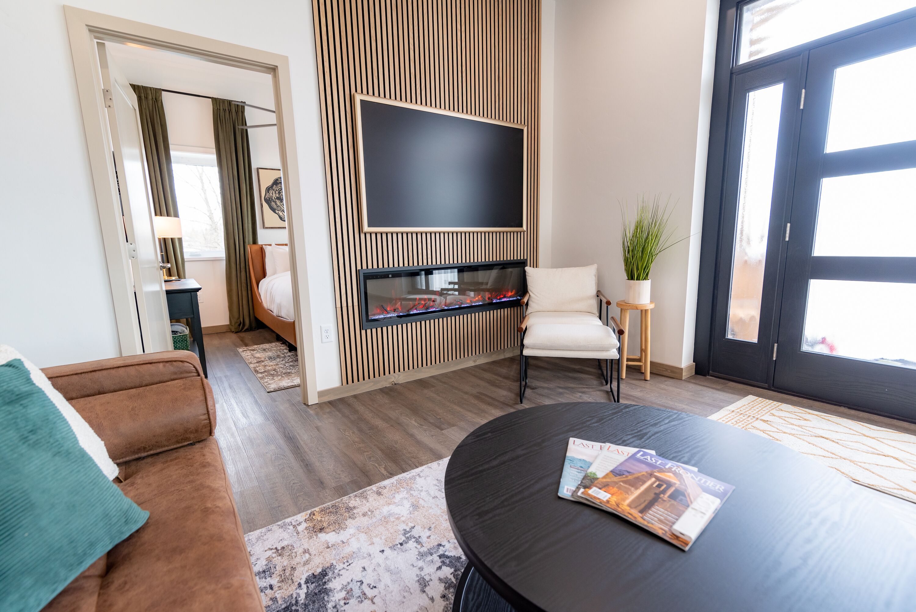 A modern living room with a flat-screen TV mounted on a wooden panel wall, a brown sofa, a round coffee table with a magazine, and a door with glass panels.