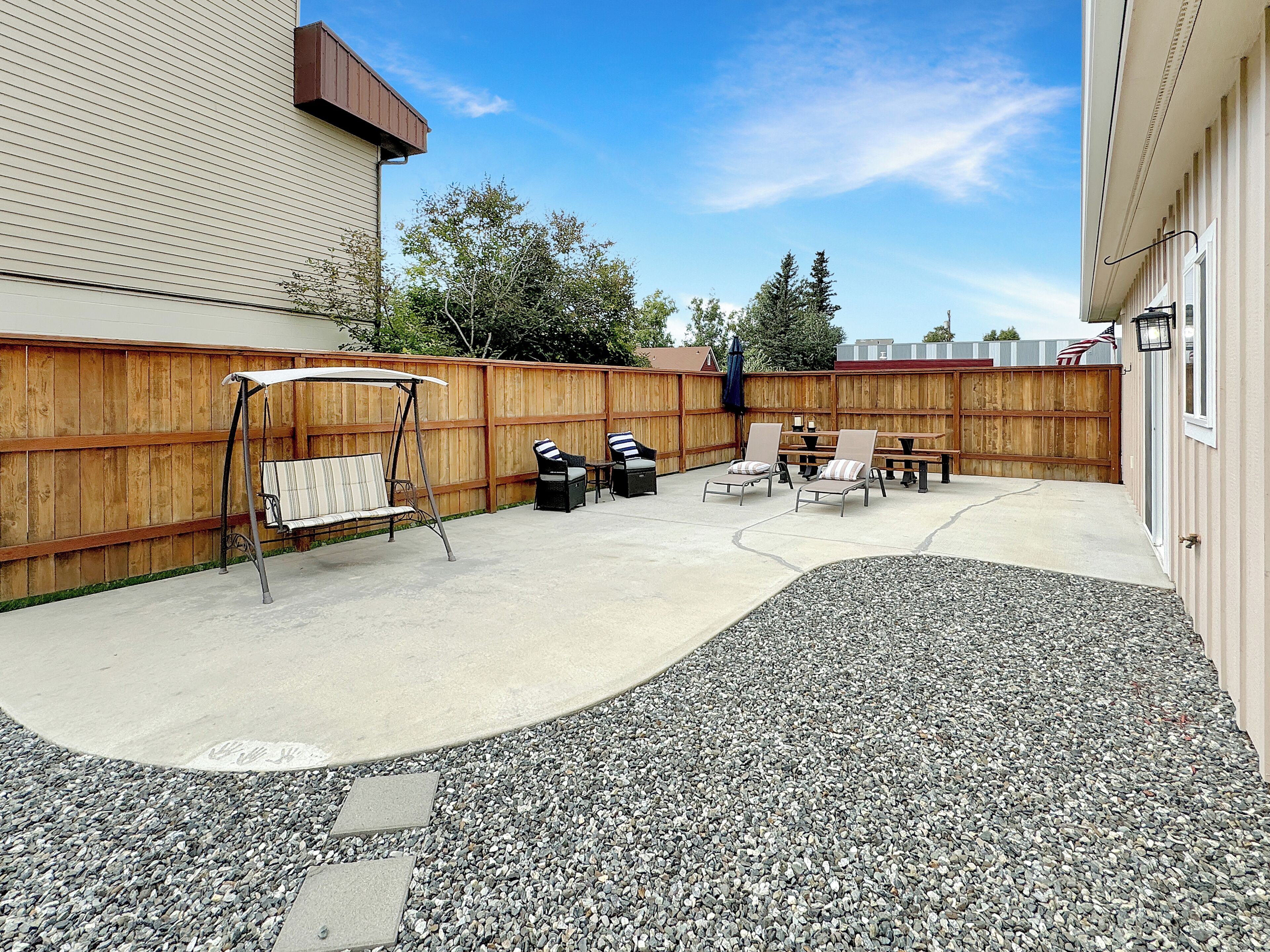 A backyard patio with a concrete floor, a wooden fence, and outdoor furniture including a swing set, chairs, and a table.