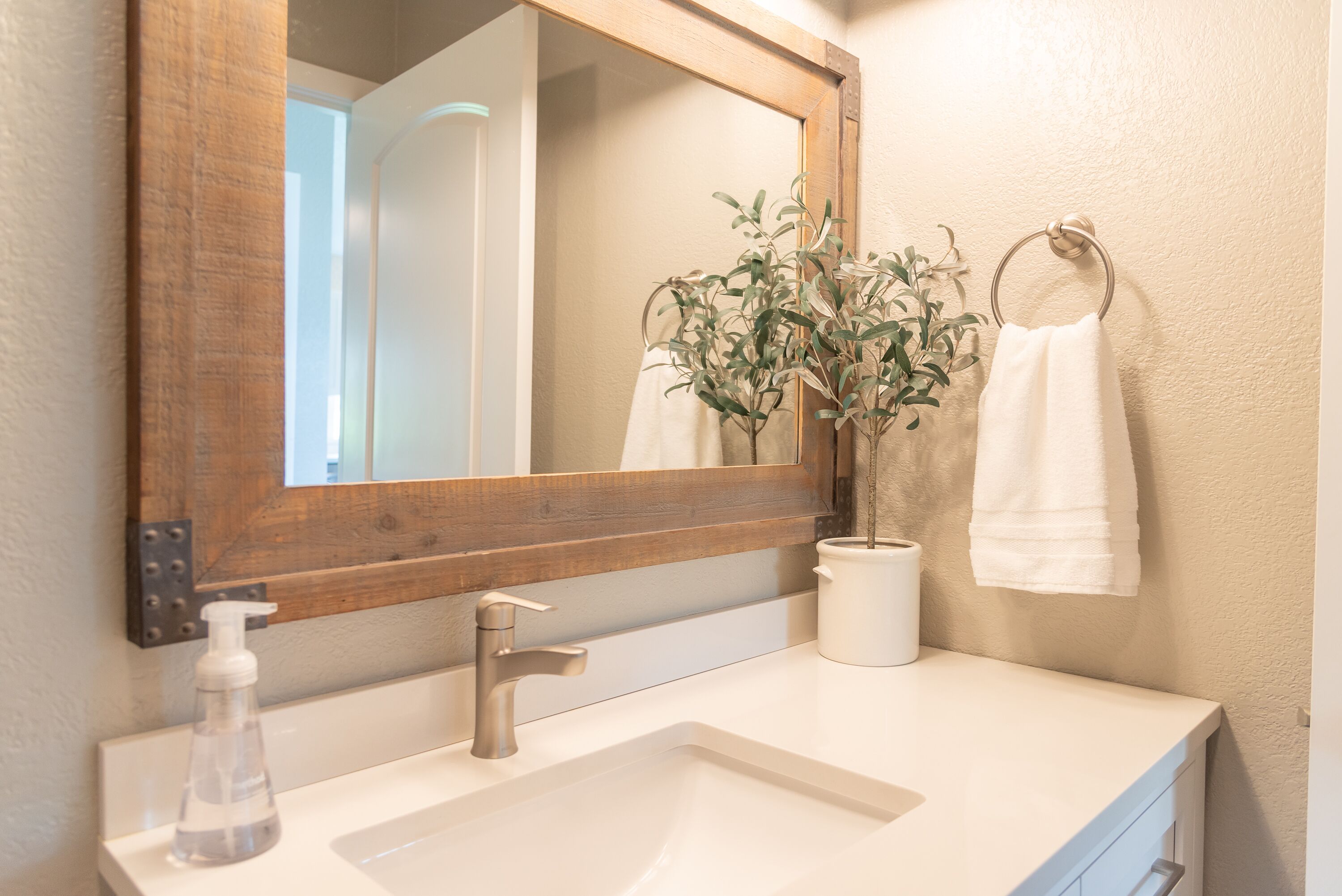 Un baño con espejo de marco de madera, lavabo blanco, dispensador de jabón y planta en maceta.