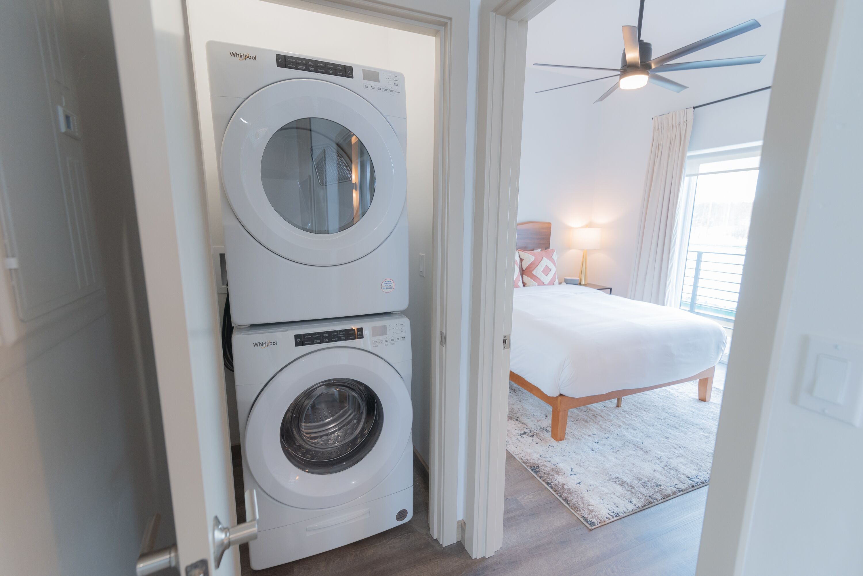 A laundry room with two stacked Whirlpool washers and dryers, a bed with a white comforter, a ceiling fan, and a window with curtains.