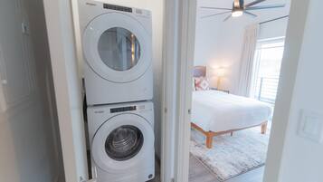 A laundry room with two stacked Whirlpool washers and dryers, a bed with a white comforter, a ceiling fan, and a window with curtains.