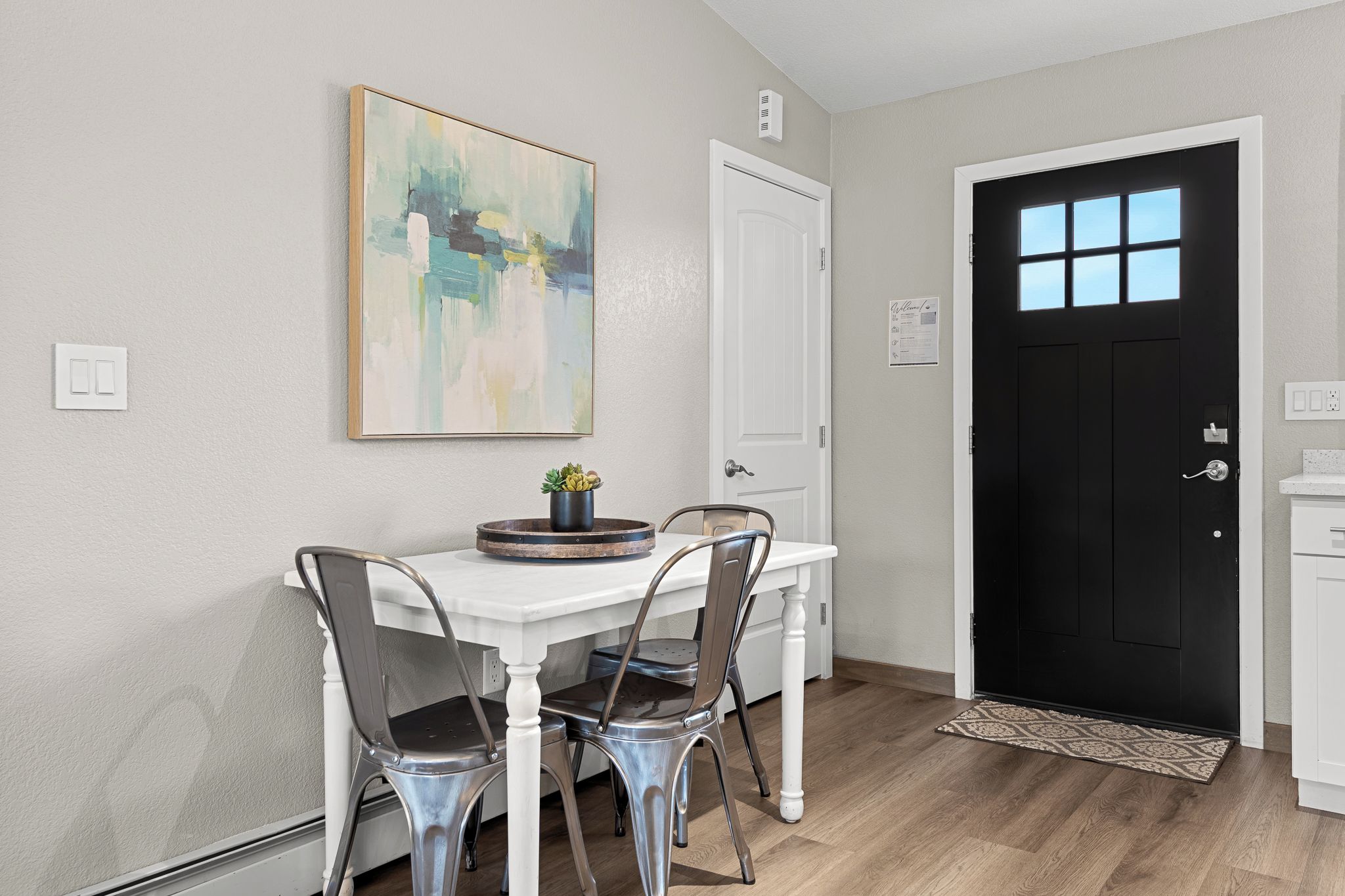 Uma área de refeições com uma mesa branca, duas cadeiras, um quadro e uma porta preta.