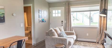 A living room with a beige sofa, a wooden table, and a small stool. There is a window with curtains, a door, and a ceiling light fixture.