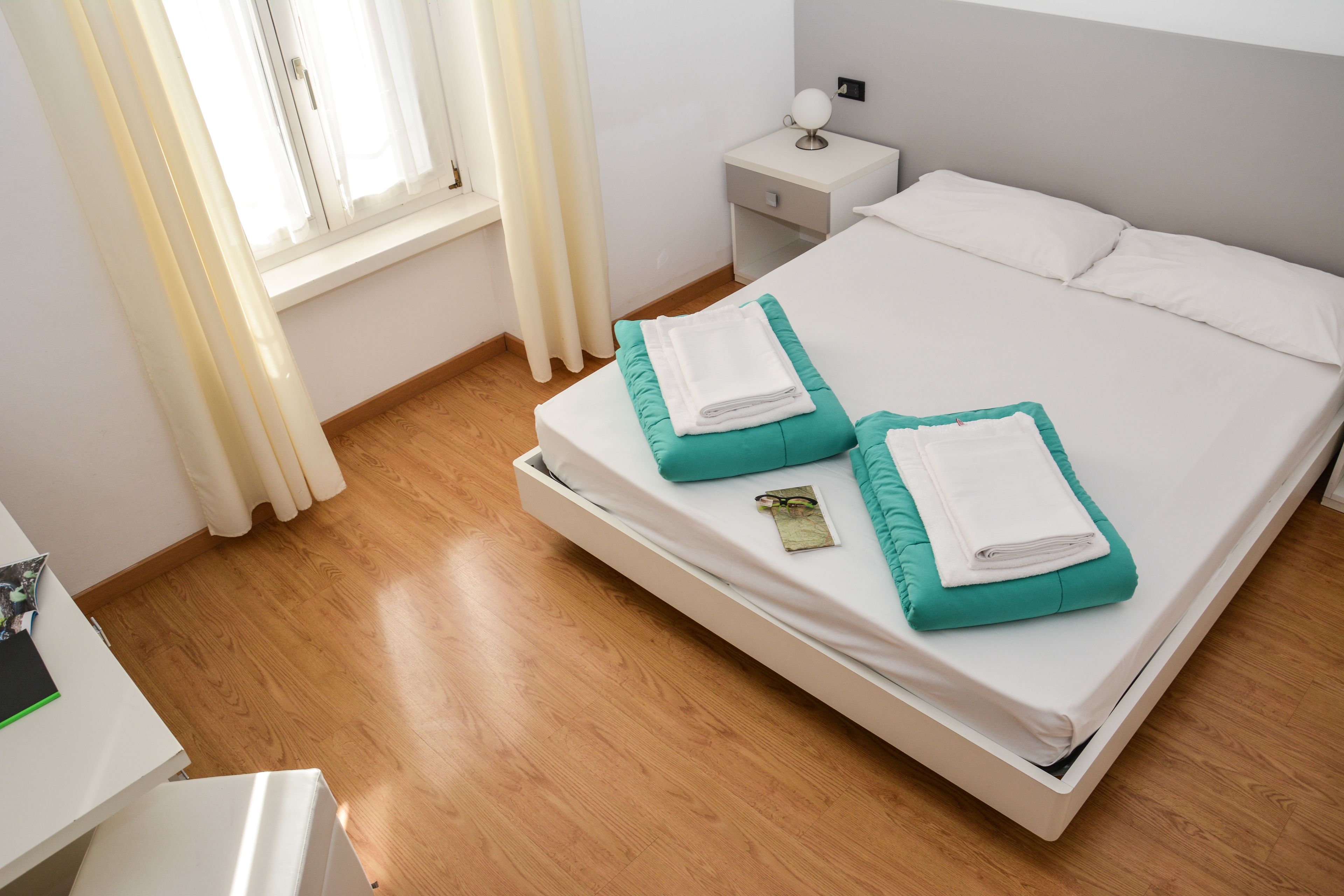 Una cama bien hecha con ropa de cama blanca y verde azulada, una mesita de noche con lámpara y una ventana con cortinas.