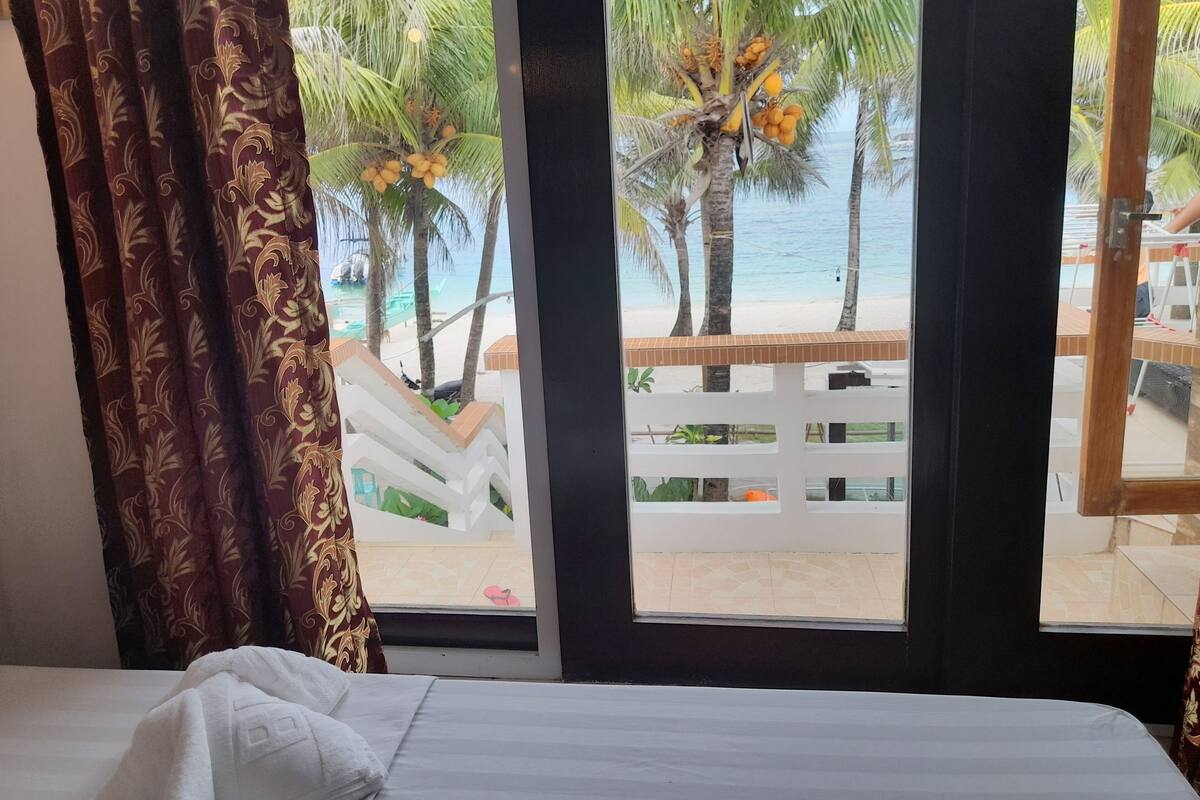 A hotel room with a bed, a window with curtains, and a view of a beach with palm trees and a pool.