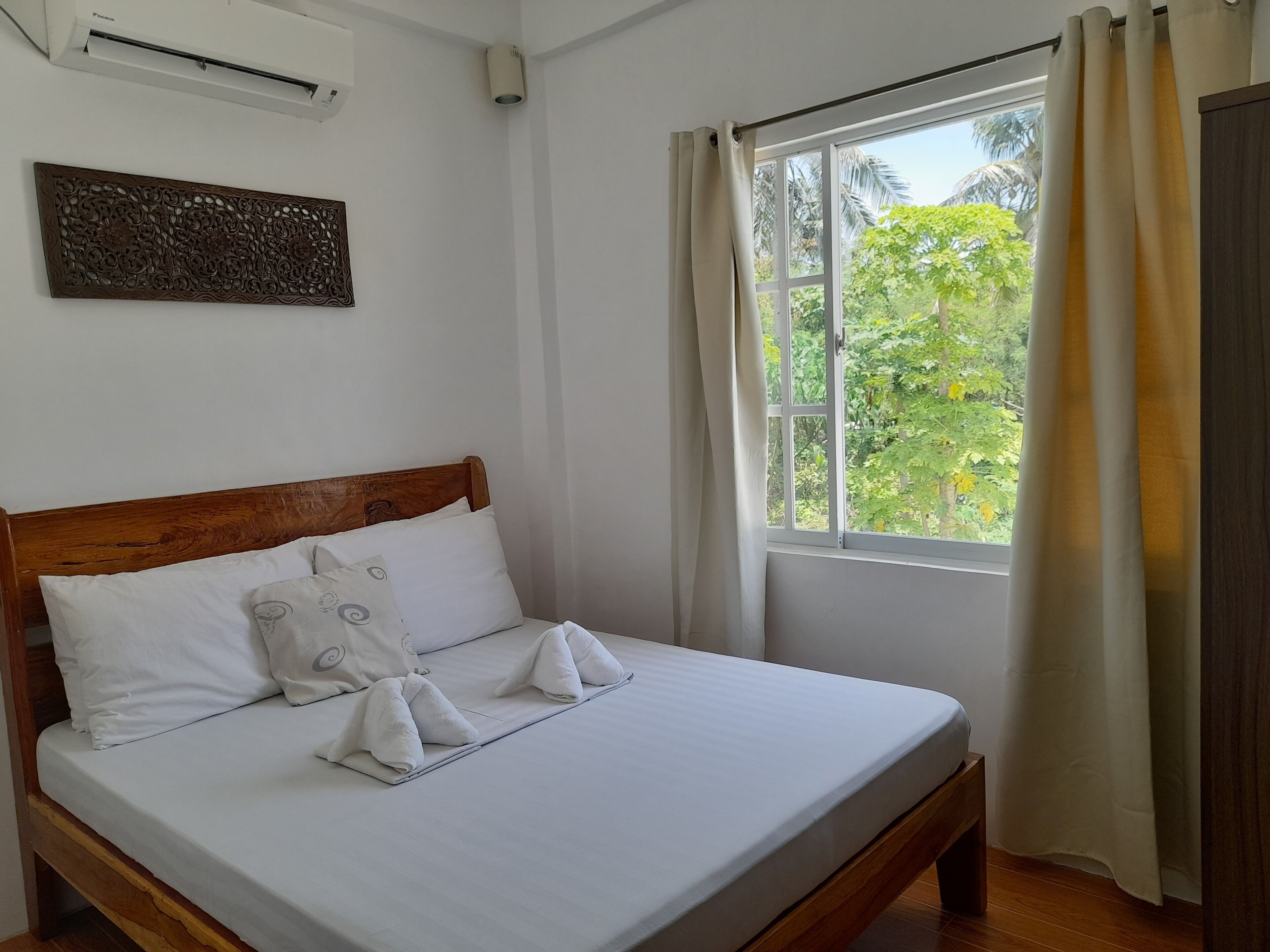 Una cama bien hecha con sábanas y almohadas blancas, cabecera de madera, aire acondicionado de pared y una ventana con cortinas.