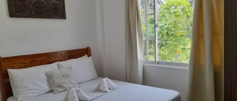 A neatly made bed with white linens and pillows, a wooden headboard, a wall-mounted air conditioner, and a window with curtains.