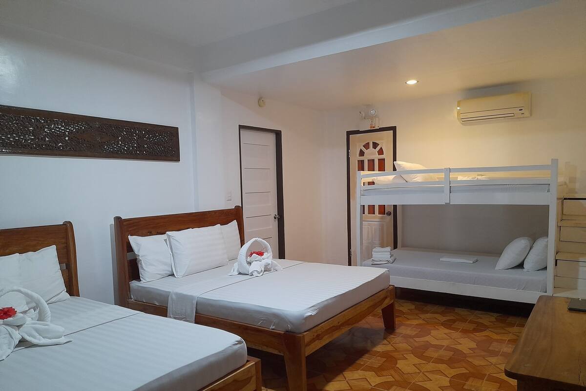 A hotel room with two single beds, a bunk bed, a wooden table, and an air conditioning unit.