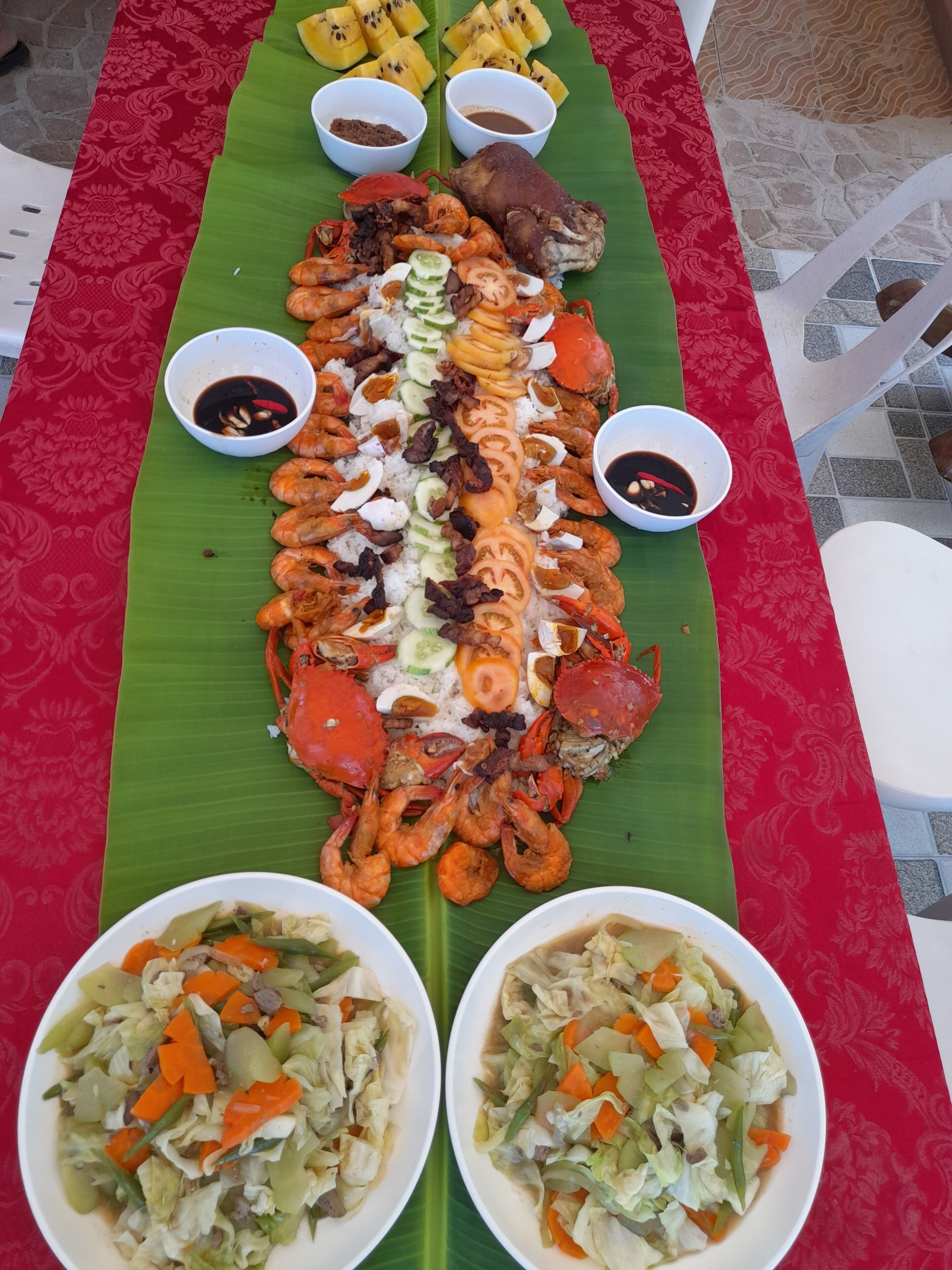 Un gran plato de mariscos y verduras sobre una hoja de plátano, acompañado de recipientes con salsas para mojar.