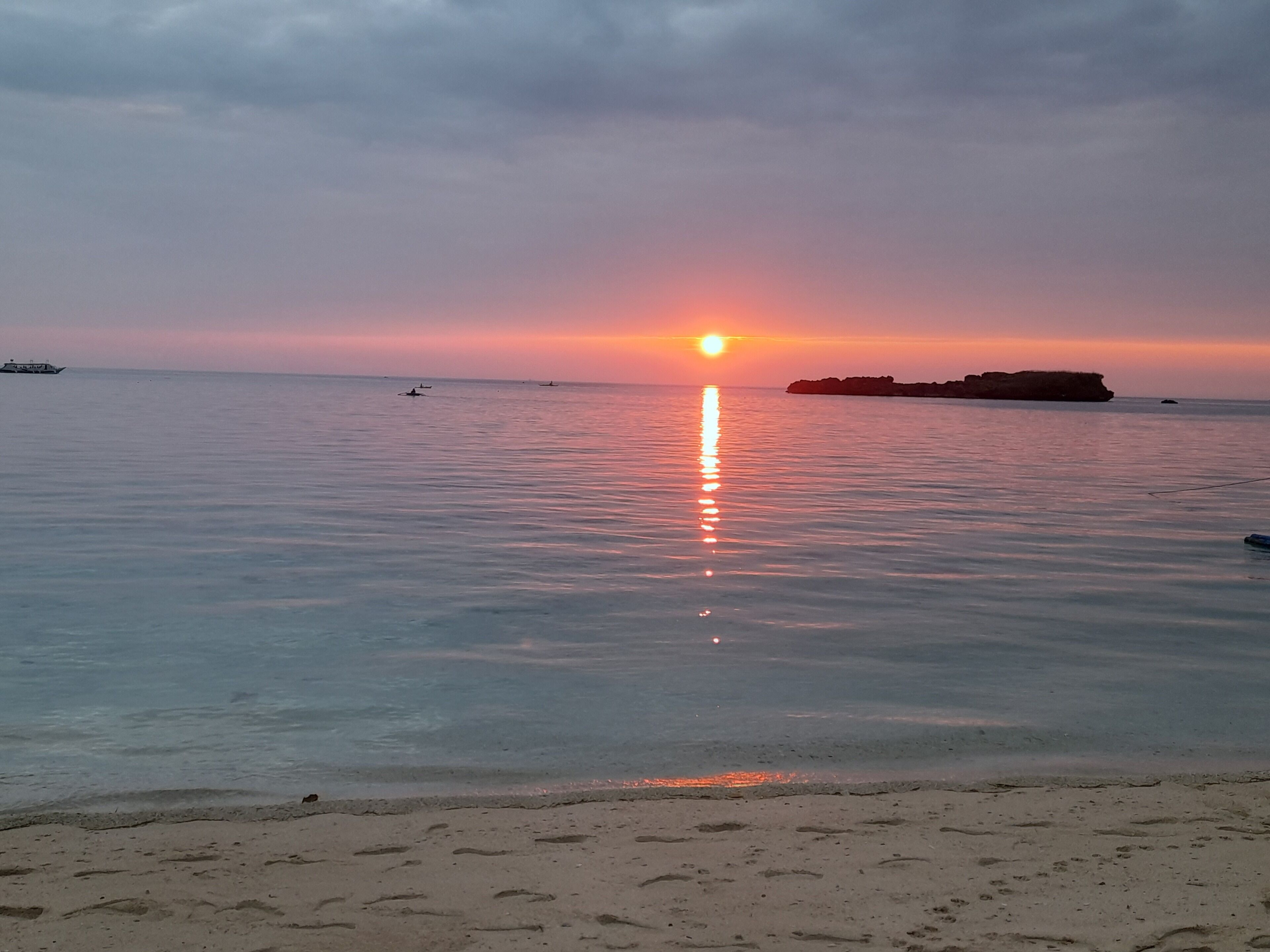 Una playa con aguas tranquilas que reflejan el atardecer, un promontorio rocoso a lo lejos y algunas embarcaciones.