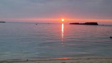 Una playa con aguas tranquilas que reflejan el atardecer, un promontorio rocoso a lo lejos y algunas embarcaciones.