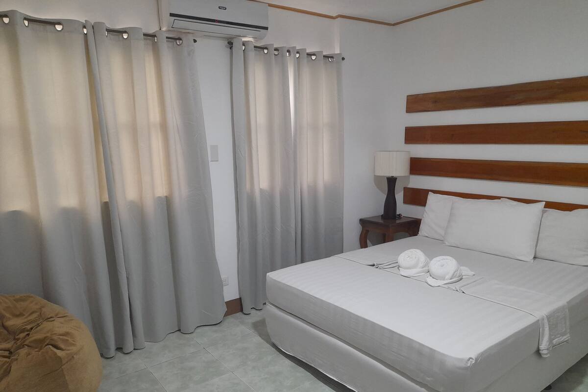A hotel room with a bed, a nightstand with a lamp, a wooden headboard, and a large window with curtains.