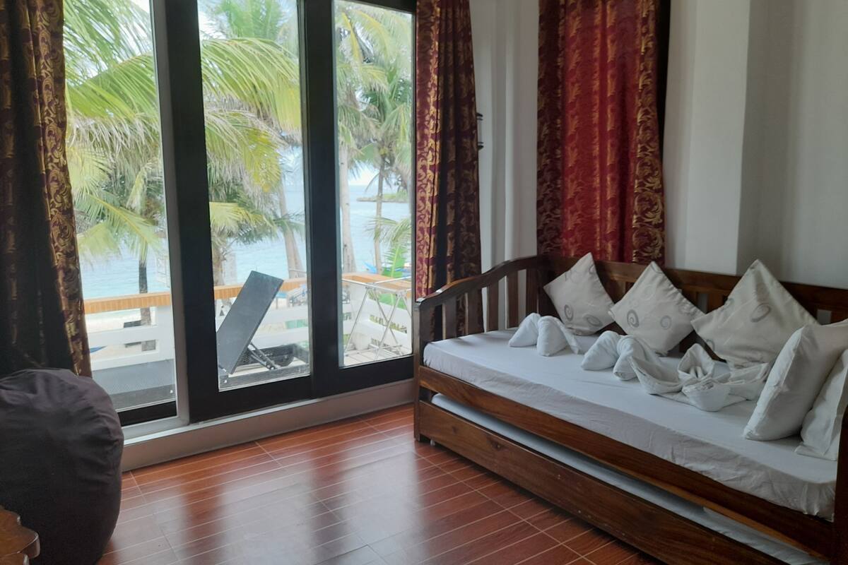 A room with a wooden floor, a bed with white bedding, and a view of palm trees and a beach through large windows.