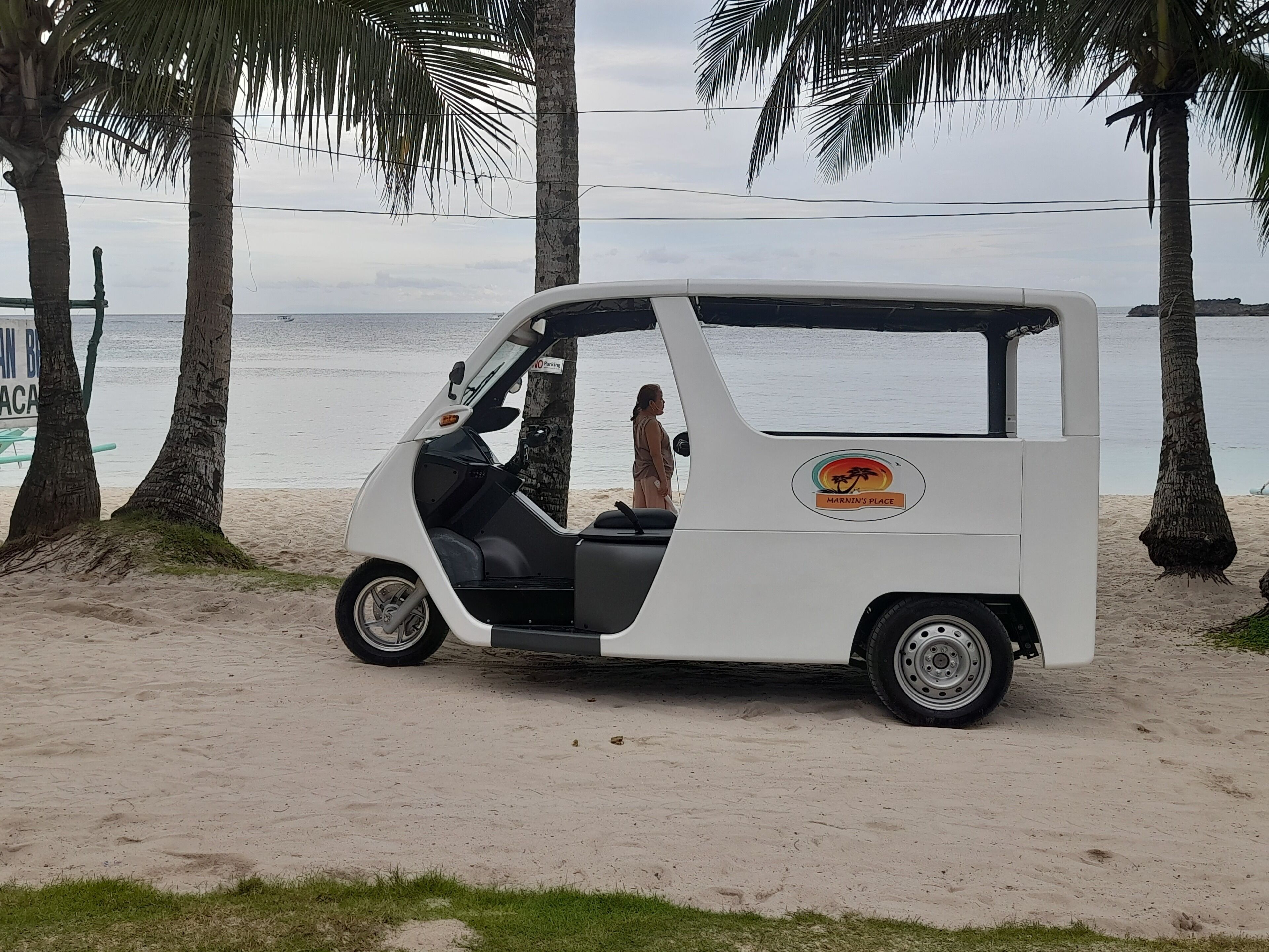 Un vehículo blanco de tres ruedas estacionado en una playa arenosa, con palmeras y una masa de agua al fondo.