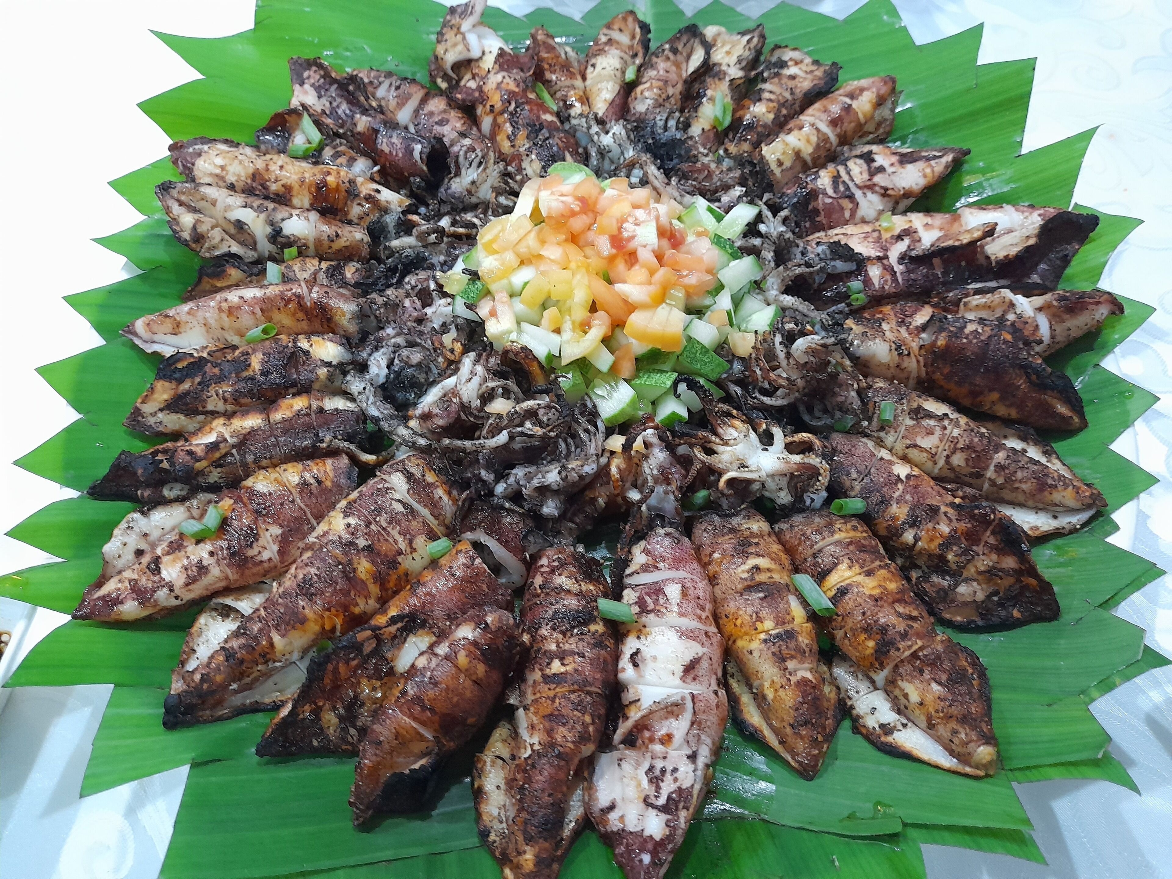 A platter of grilled octopus arranged in a circular pattern on a banana leaf, with a diced mango and cucumber salsa in the center.