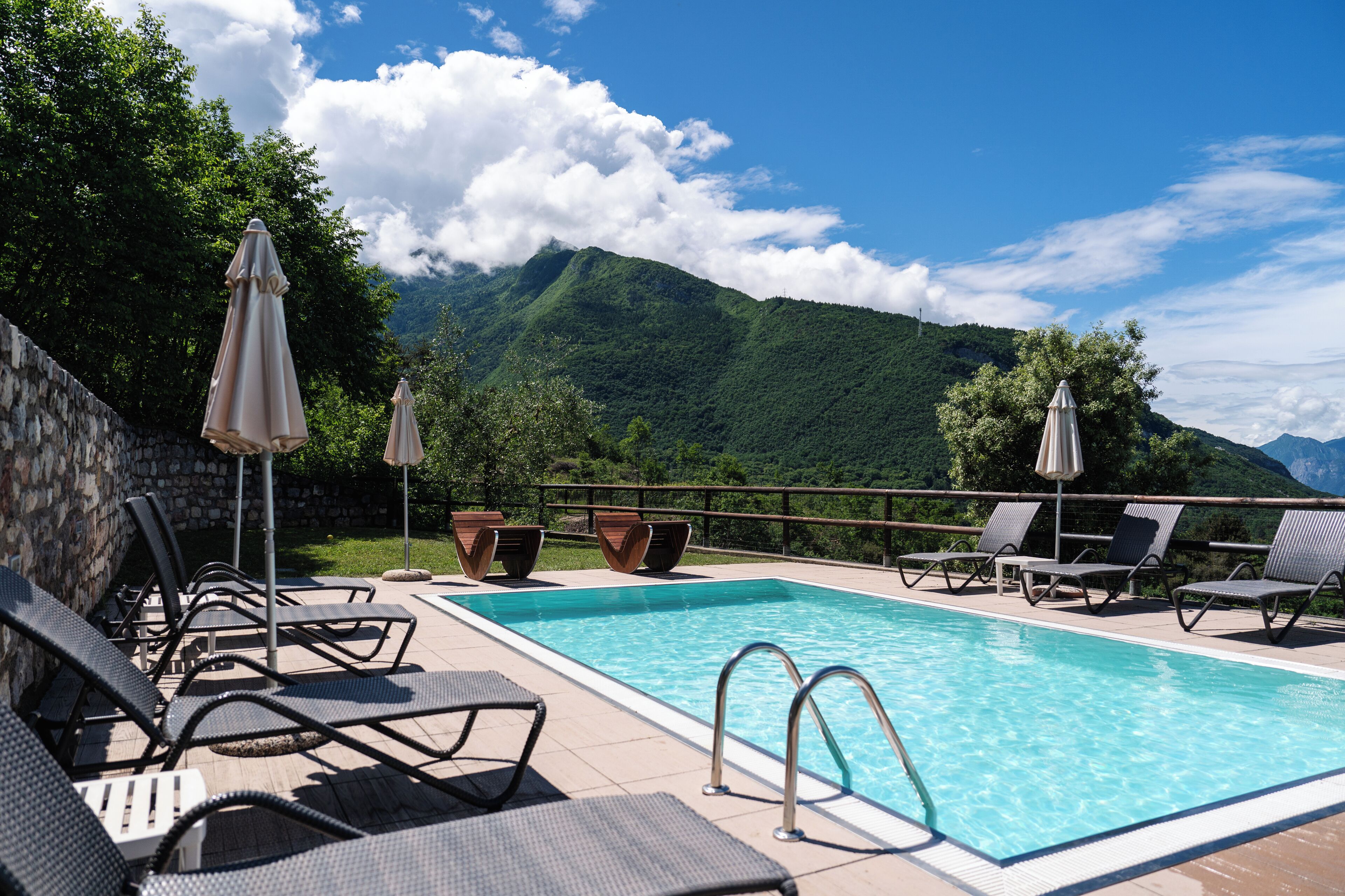 A swimming pool with lounge chairs and umbrellas, surrounded by a stone wall and lush greenery.