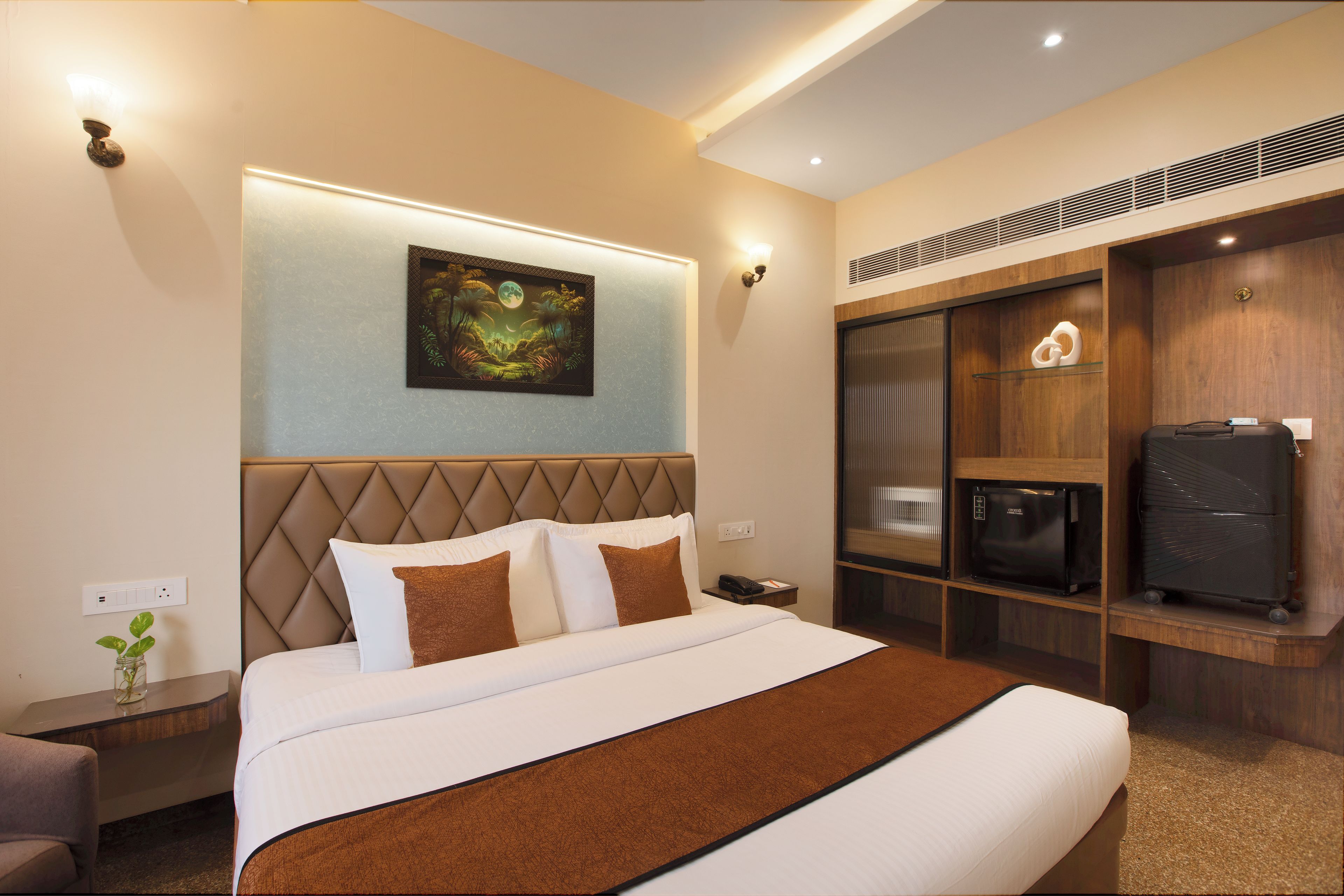 A hotel room with a bed, a nightstand, a wall-mounted painting, a wardrobe, and a minibar.